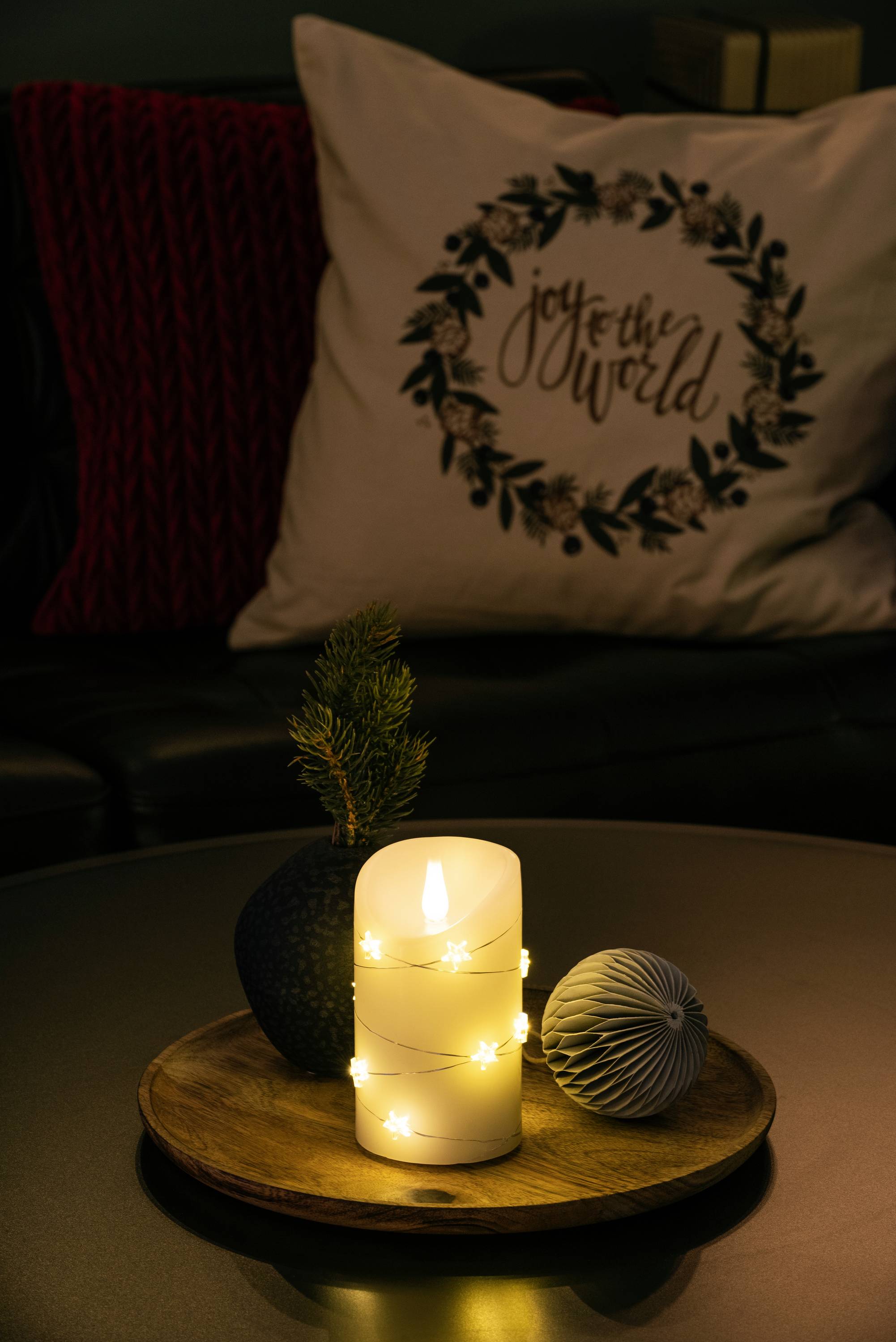 Een brandende kaars met sterrenlichjes op een houten dienblad naast decoraties. Op de achtergrond een kussen met 'vreugde voor de wereld'.