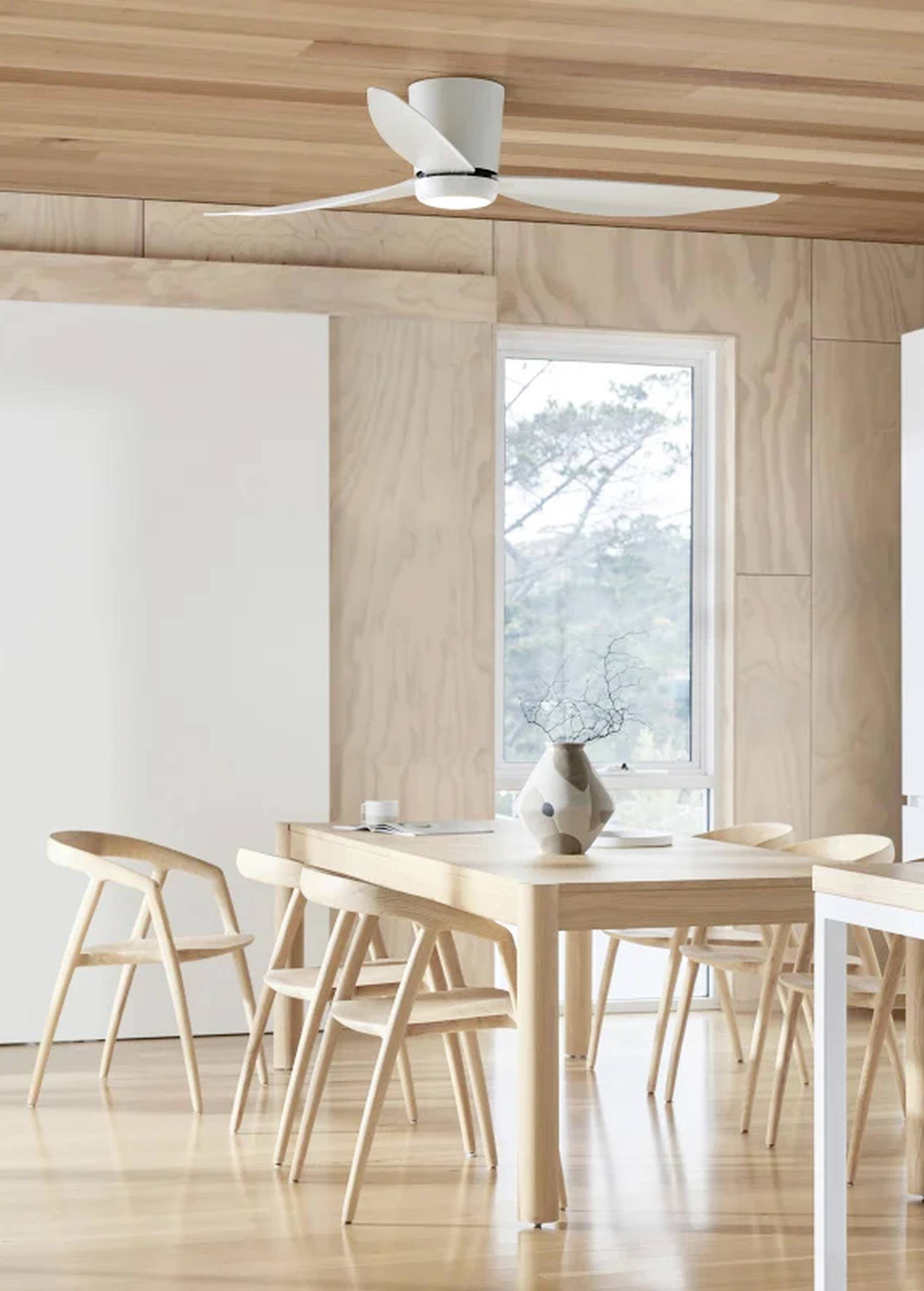 Lichte eetkamer met houten tafel en stoelen, vaas met tak op tafel, grote ventilator aan het houten plafond, raam op de achtergrond.