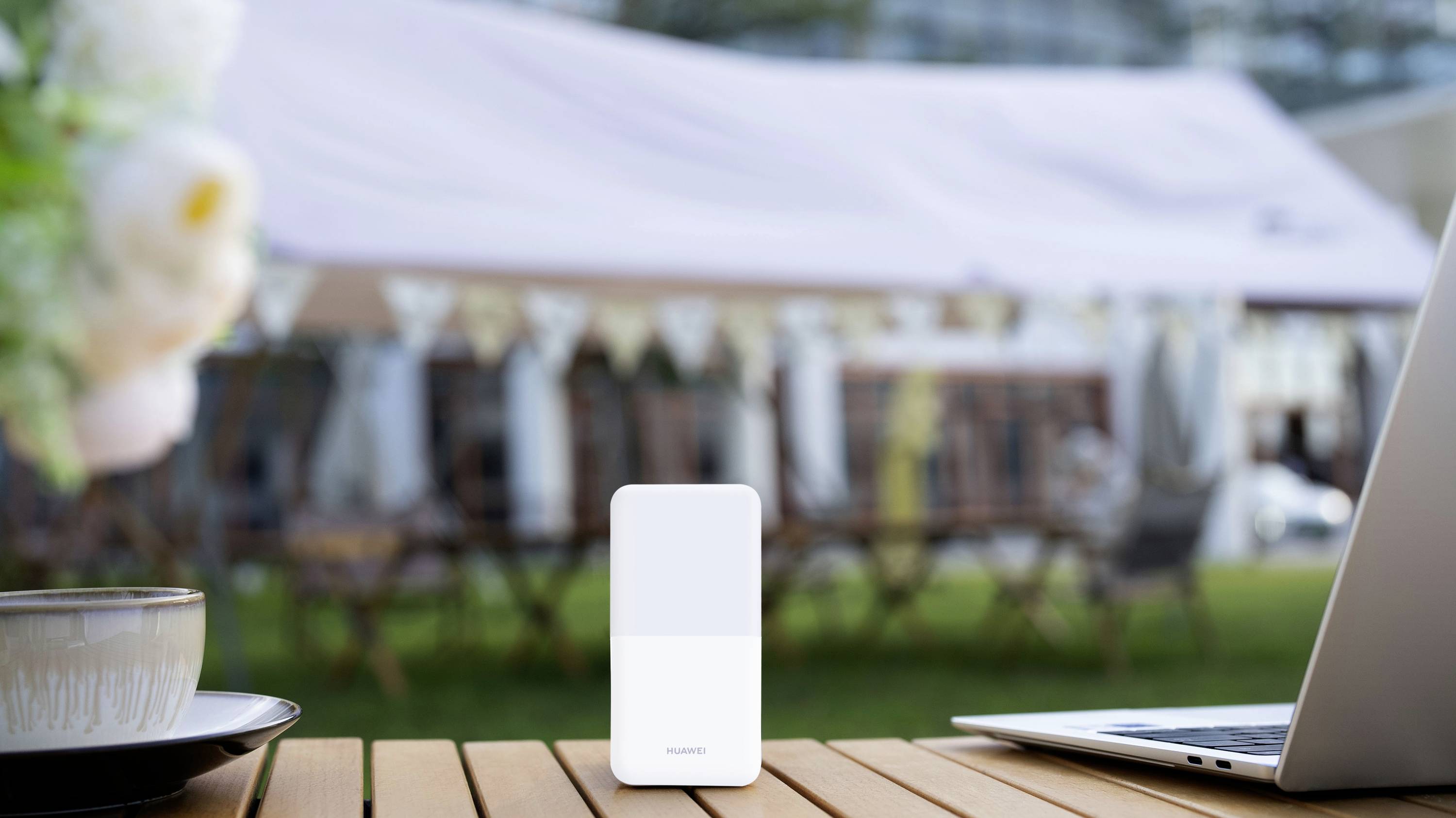 Een mobiele wifi-router staat op een tafel buiten, naast een kopje en een laptop; op de achtergrond een feesttent.