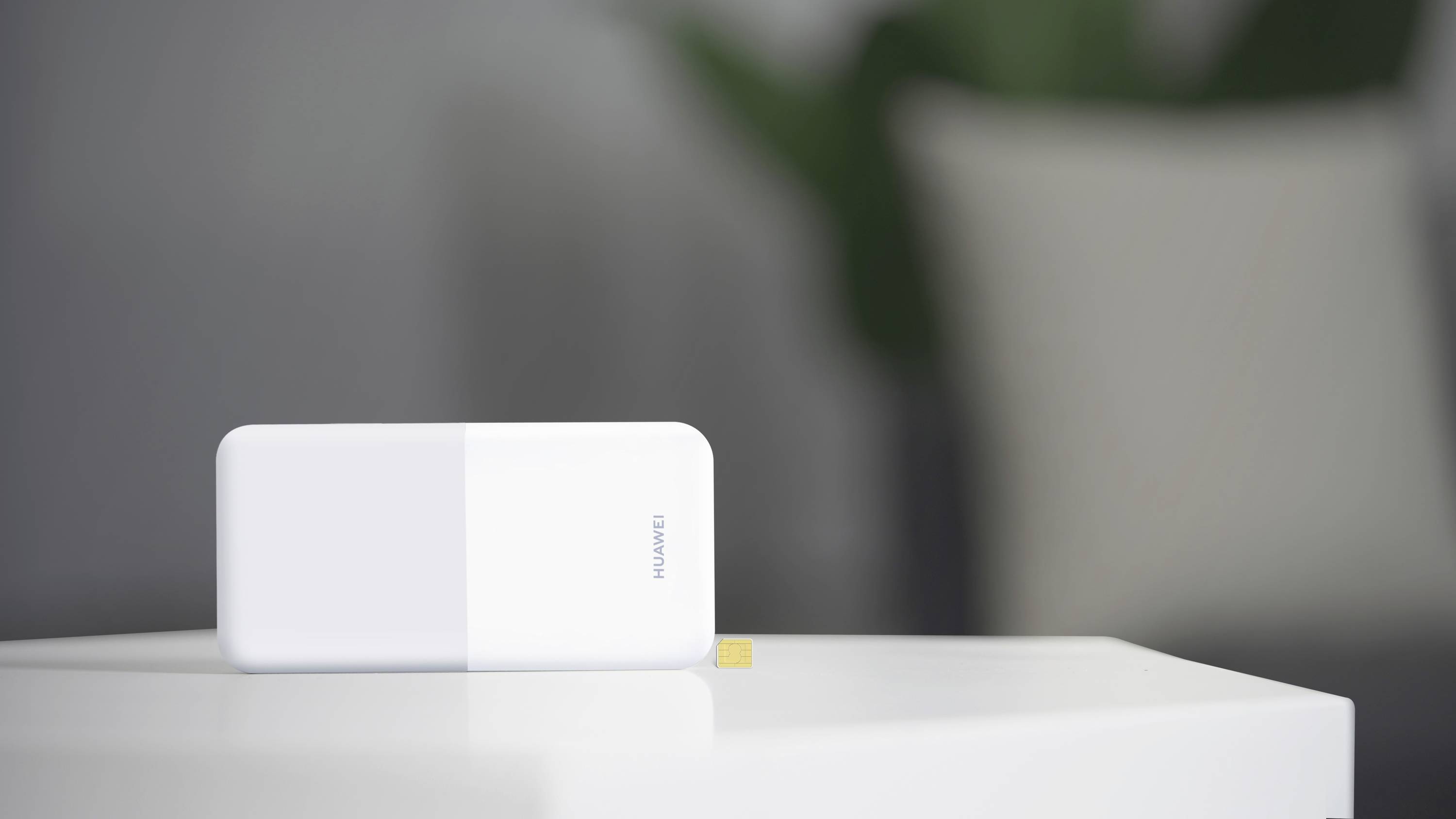 Een witte, rechthoekige Huawei-wifi-router staat op een witte tafel. De achtergrond is onscherp met groene plantenonderdelen.