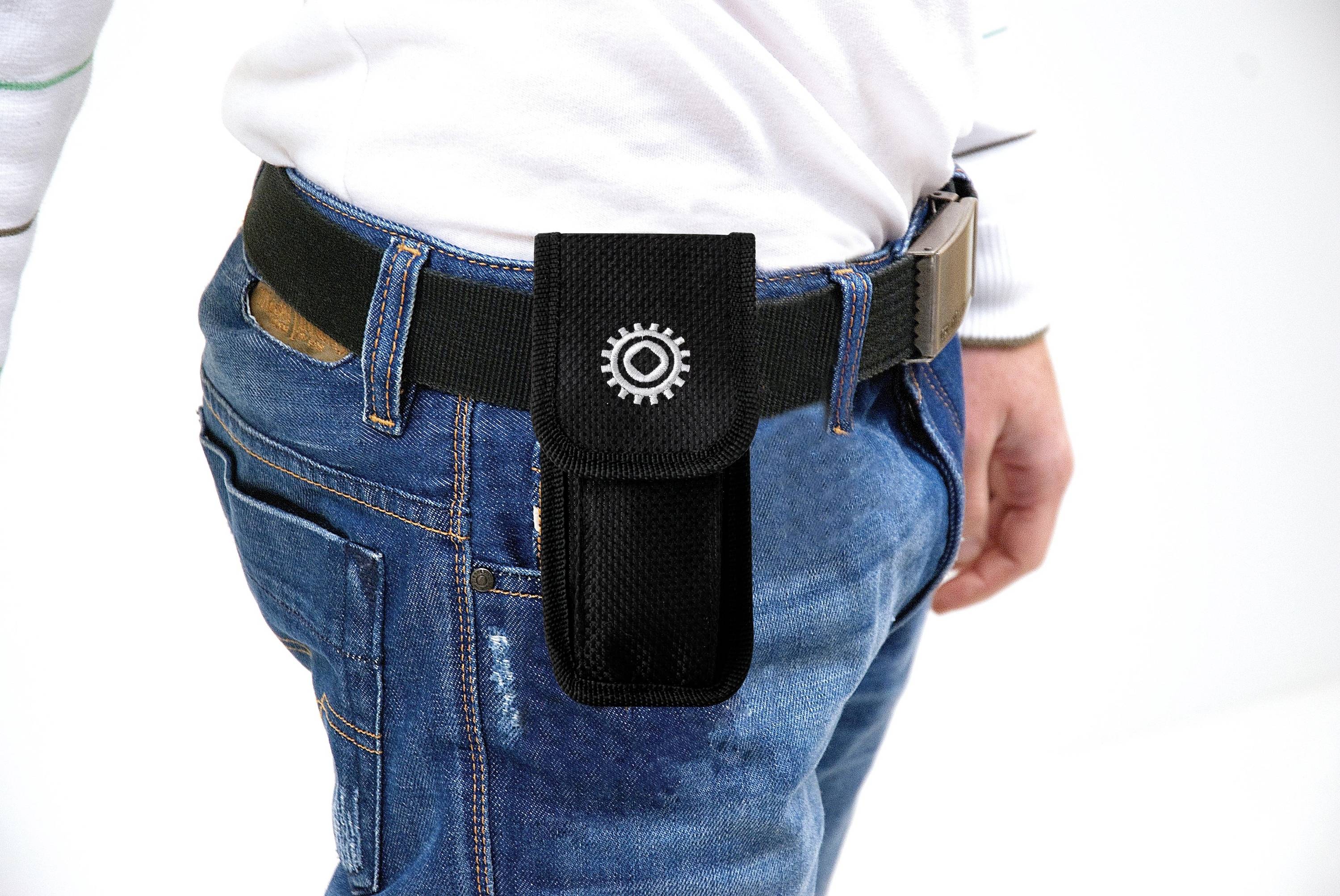 Een persoon draagt een zwarte etui met een tandwielontwerp aan de riem, gedragen over een blauwe spijkerbroek.