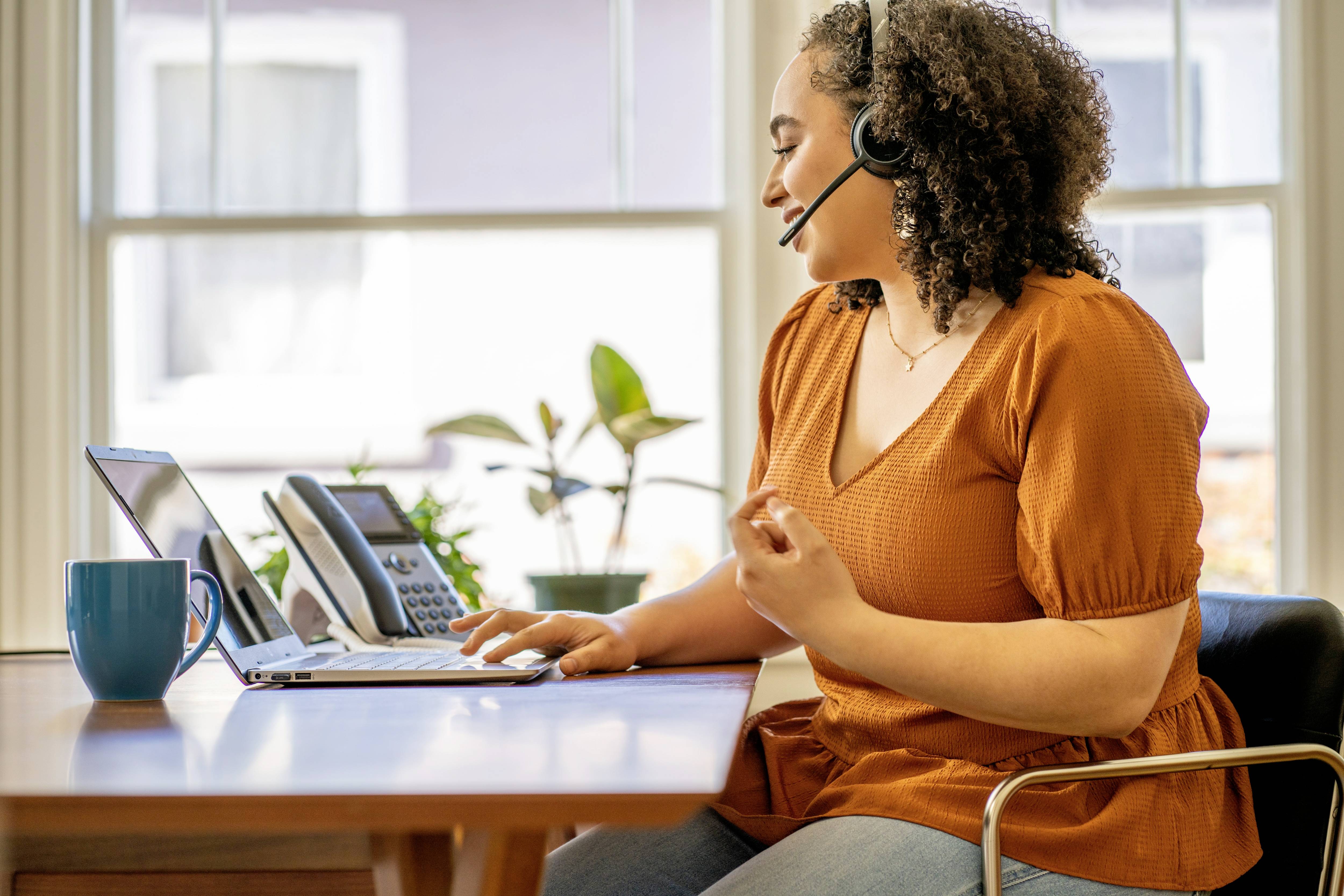 Een vrouw met een headset werkt aan een laptop. Een telefoon en een kopje staan op tafel. Op de achtergrond zijn ramen en planten.