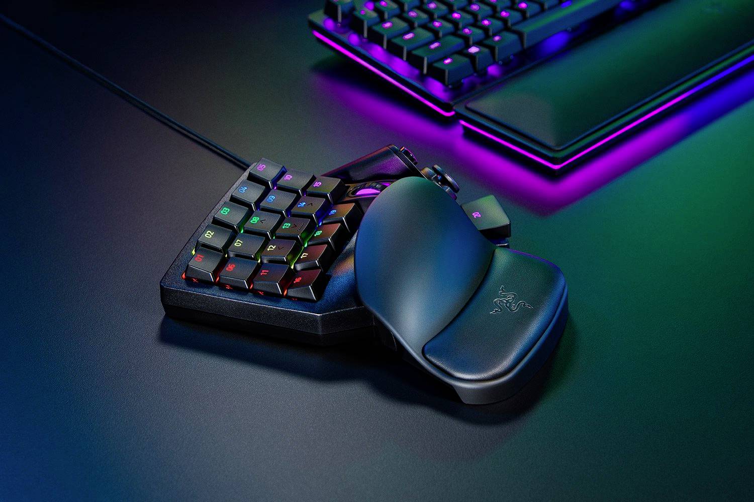 Een compact, verlicht gaming-keypad met een ergonomische handsteun, omgeven door een verlichte gaming-toetsenbord op een bureau.