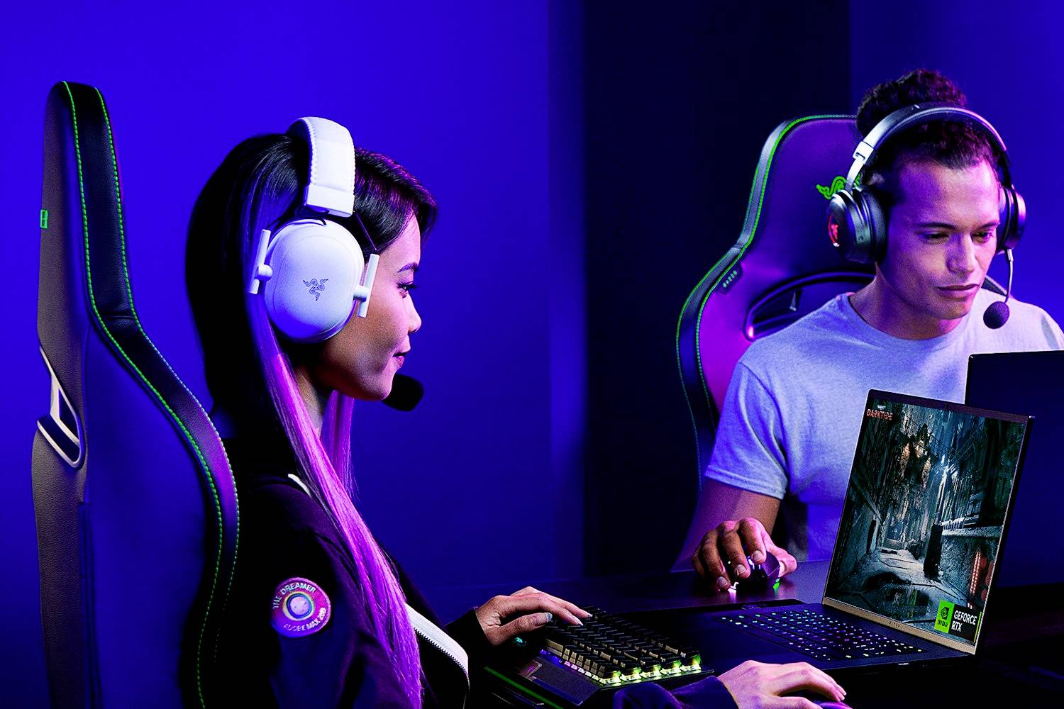 Twee personen spelen naast elkaar videogames op computers met koptelefoons en microfoons, in een blauw verlichte ruimte.