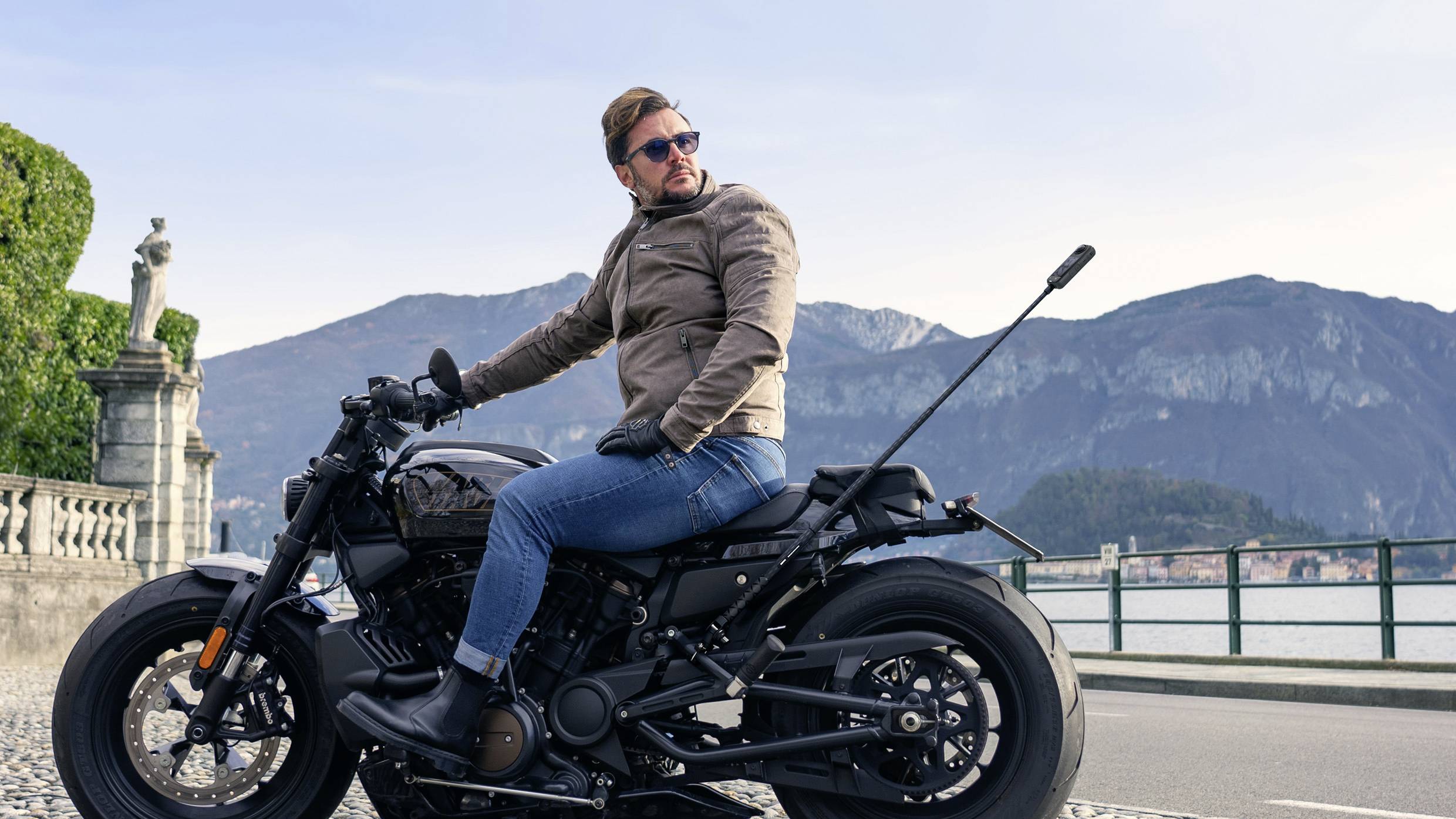 Een man zit op een motorfiets voor een berglandschap. Hij draagt een zonnebril en een bruine jas.