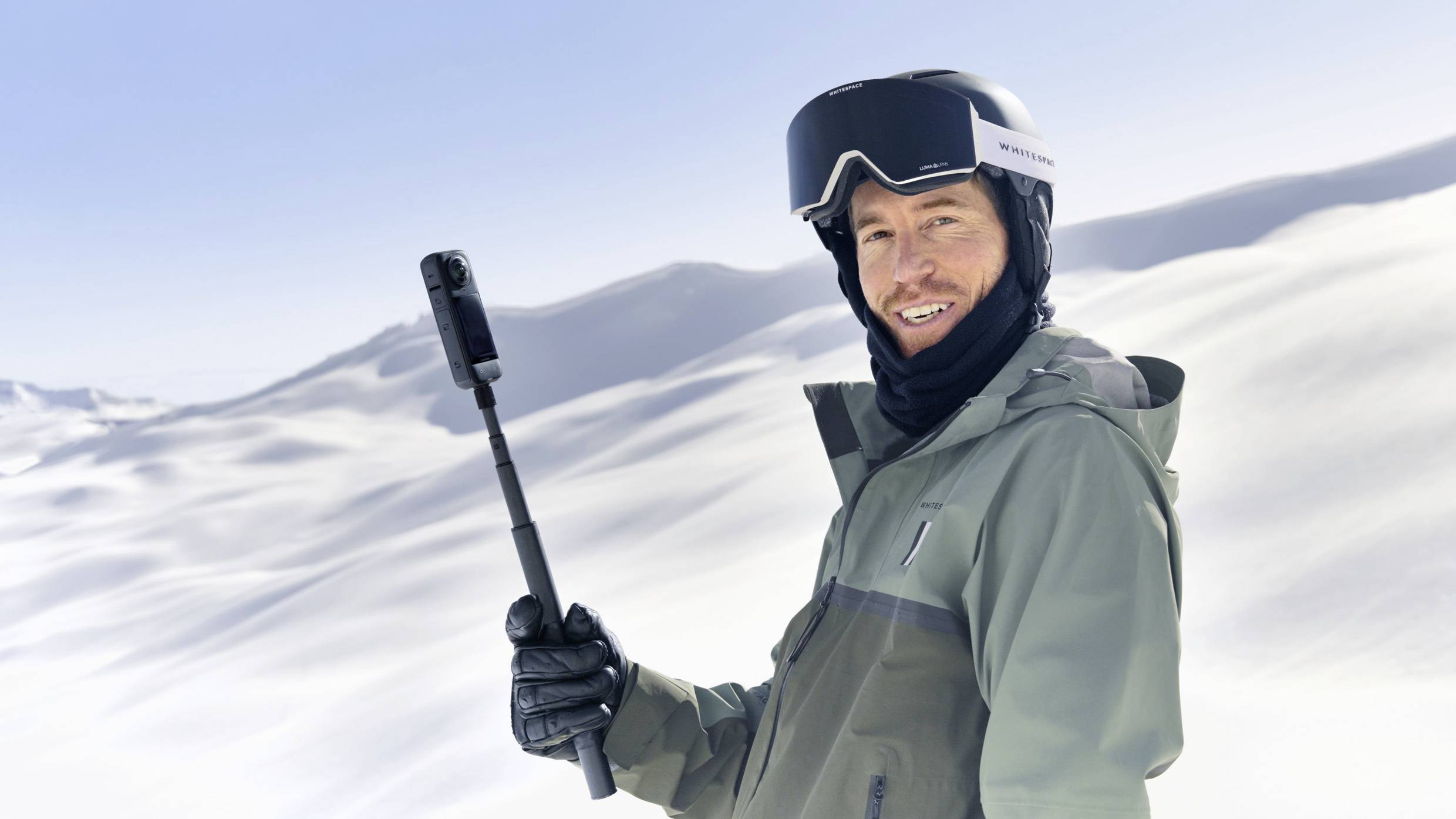 Een persoon in winterkleding houdt een selfie-stick vast en glimlacht in een met sneeuw bedekt berglandschap.
