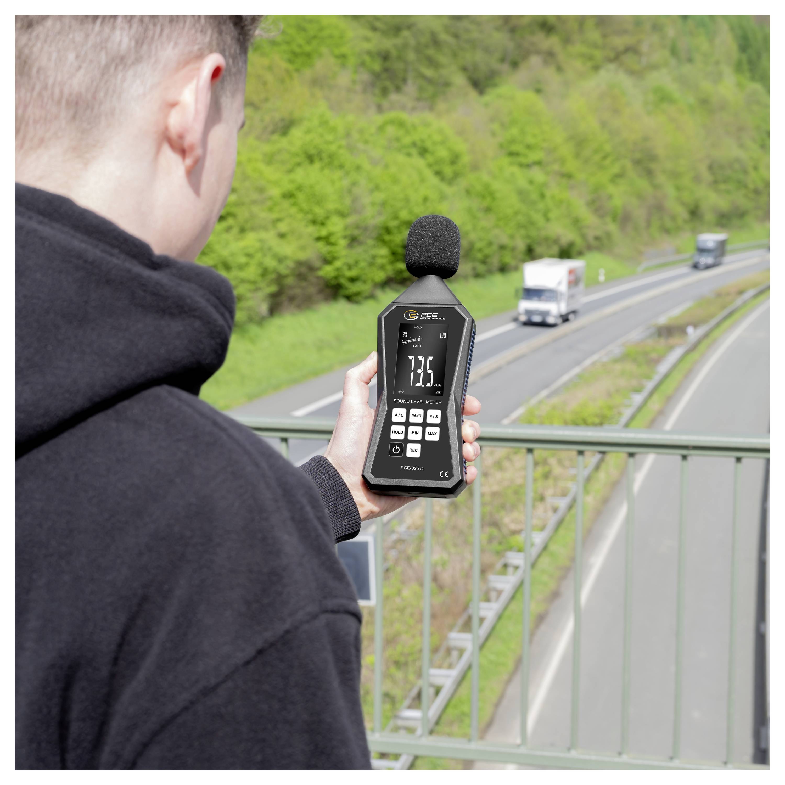Persoon die wegnoise meet met een draagbare geluidniveaumeter naast een snelweg, met een aflezing van 73,5 dB.
