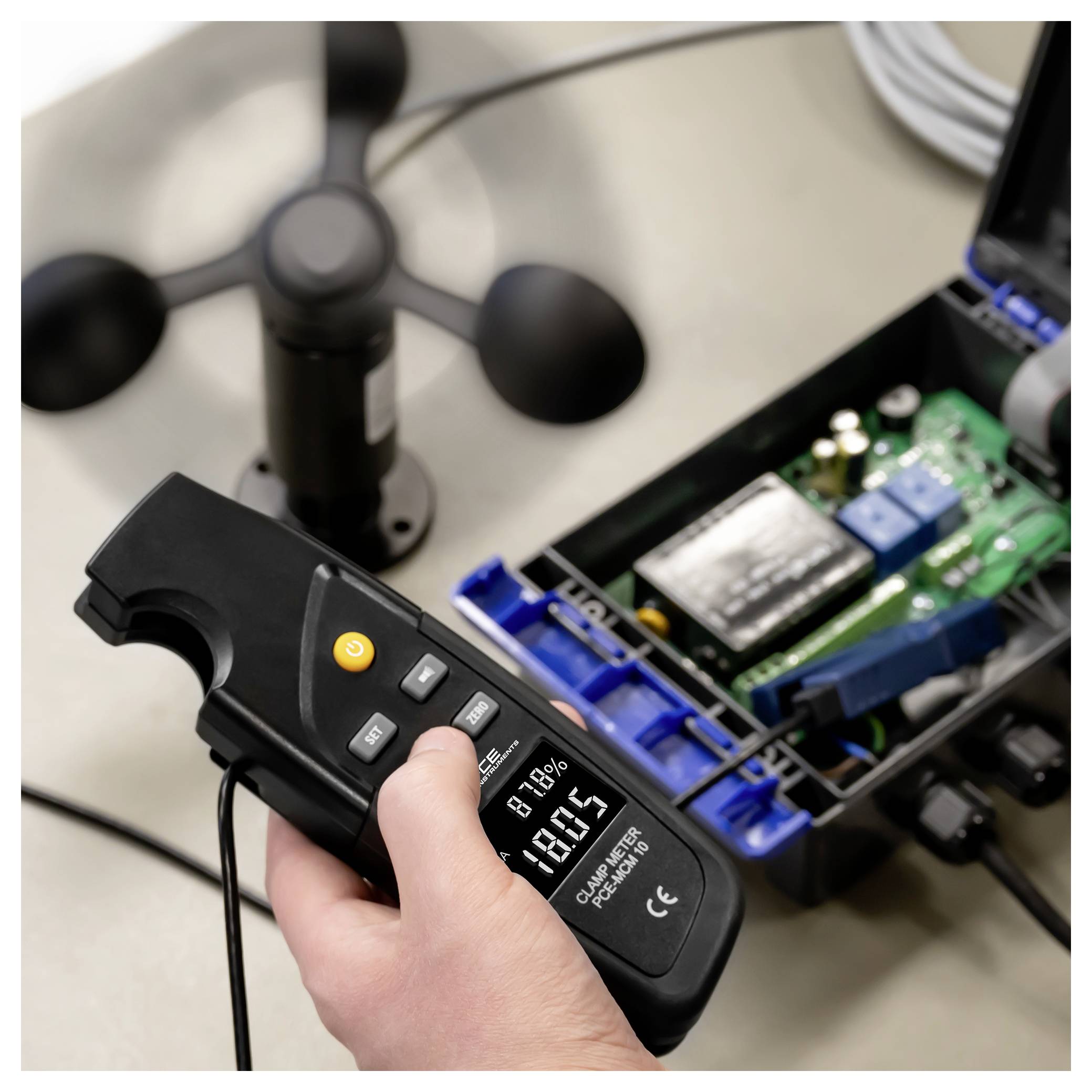 Hand die een digitale meter vasthoudt die spanning meet op een open elektronisch apparaat met een printplaat en ventilator op de achtergrond.