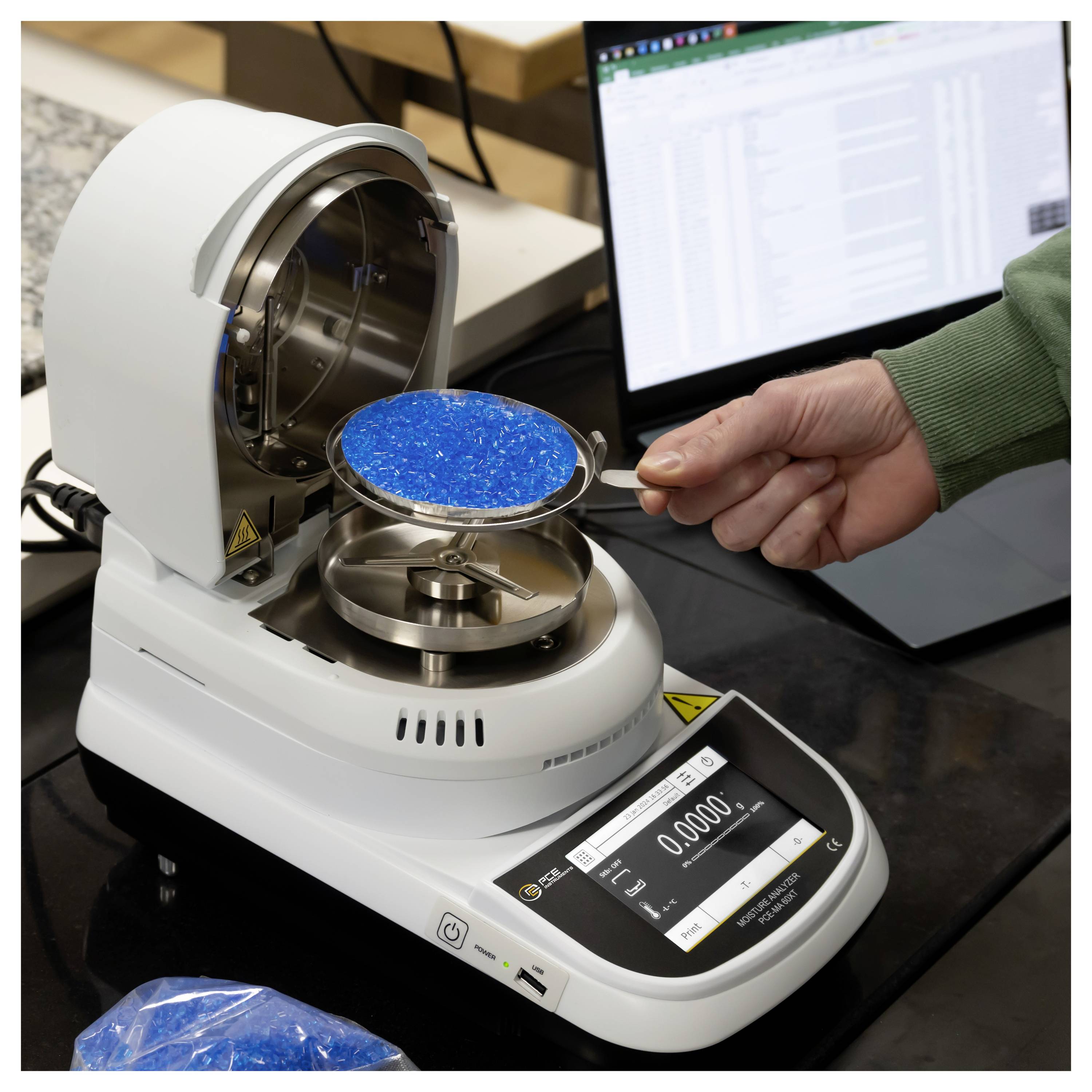 Een persoon gebruikt een laboratoriumanalytische weegschaal om een lepelvolle helder blauwe granules te wegen, met een computerscherm op de achtergrond.