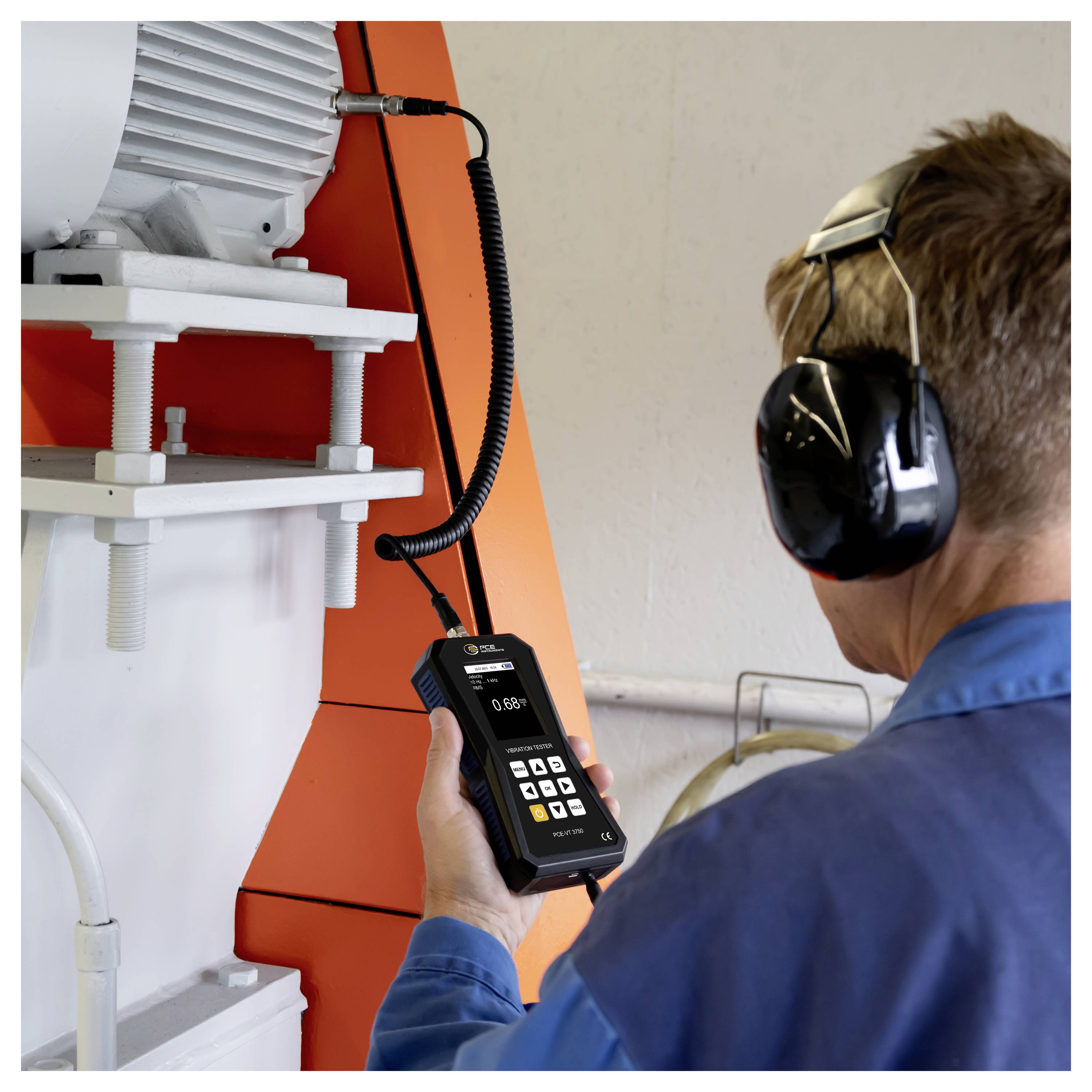 Technicus met gehoorbescherming gebruikt een handheld meter om industriële machines naast een grote motor en oranje apparatenpaneel te inspecteren.