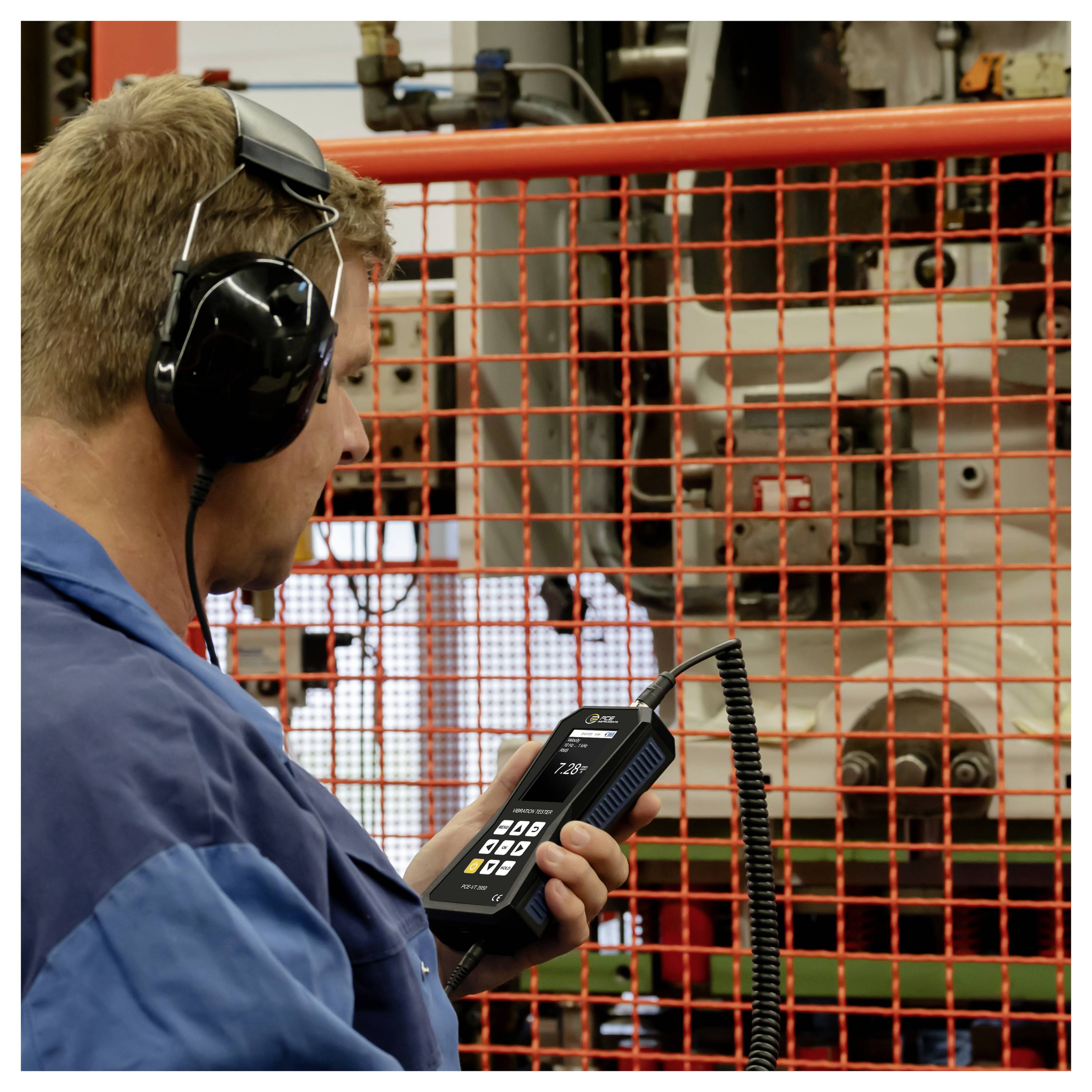Werknemer met gehoorbescherming gebruikt een handheld meetinstrument om machines achter een oranje veiligheidsbarrière te inspecteren.