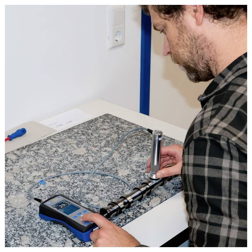 Een persoon gebruikt een apparaat om metalen staven op een werkblad te testen, met de nadruk op precisie. De scène doet denken aan een werkomgeving of laboratorium.