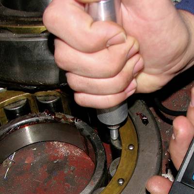 Een hand houdt een gereedschap vast en bewerkt een metalen lager. Op de achtergrond zijn tandwielen zichtbaar die wijzen op een werkplaatsomgeving.