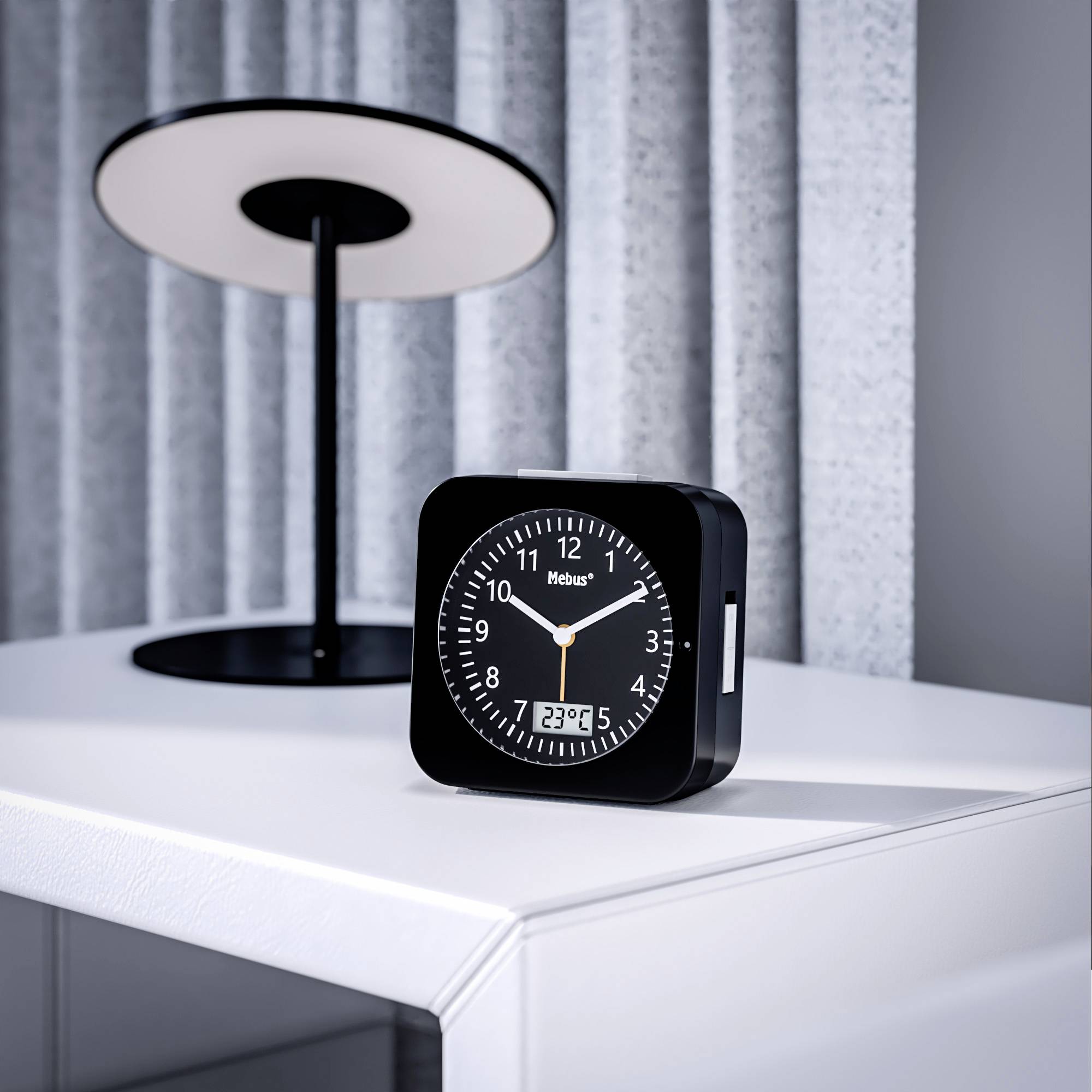 Een zwarte wekker met een wit display en digitale temperatuurweergave staat op een nachtkastje, naast een moderne lamp.