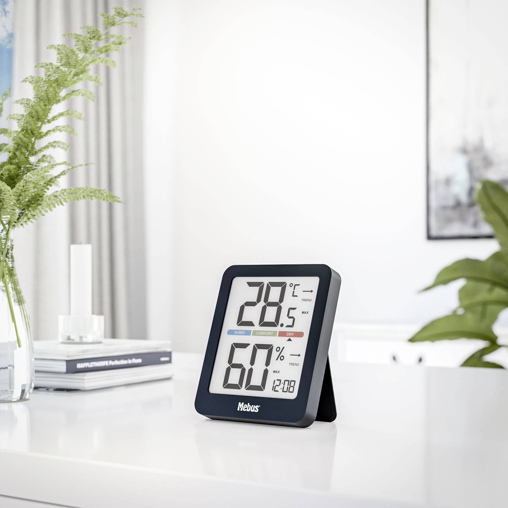 Een digitale thermo-hygrometer op een tafel toont 25,8°C en 60% luchtvochtigheid aan. Op de achtergrond zijn boeken en planten zichtbaar.