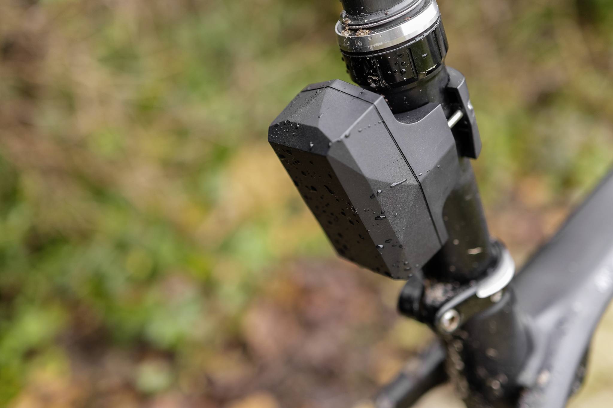 Close-up van een fietsonderdeel met een elektronisch apparaat aan het frame. Het apparaat is besproeid met regendruppels. Achtergrond onscherp, herfstachtige kleuren.