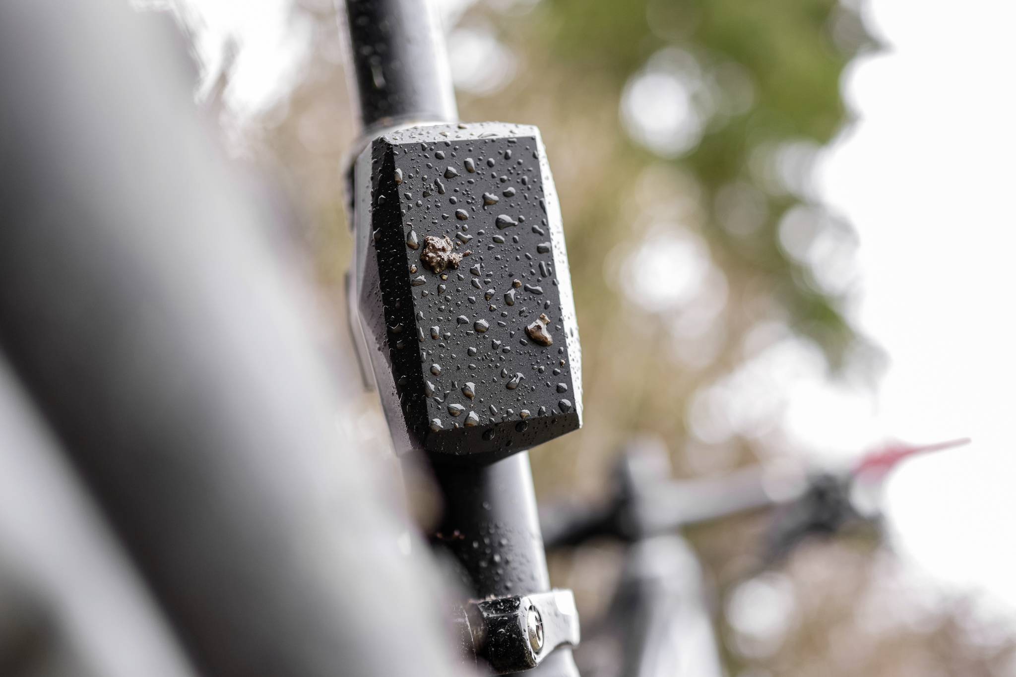 Close-up van een zwart gelakt fietskader met waterdruppels, met een onscherpe natuurlijke achtergrond.