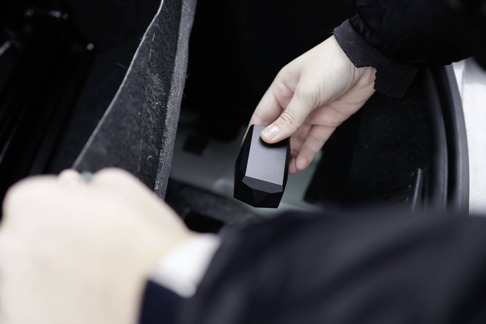 Een persoon houdt een klein, zwart, geometrisch voorwerp in de hand boven de open kofferbak van een auto.