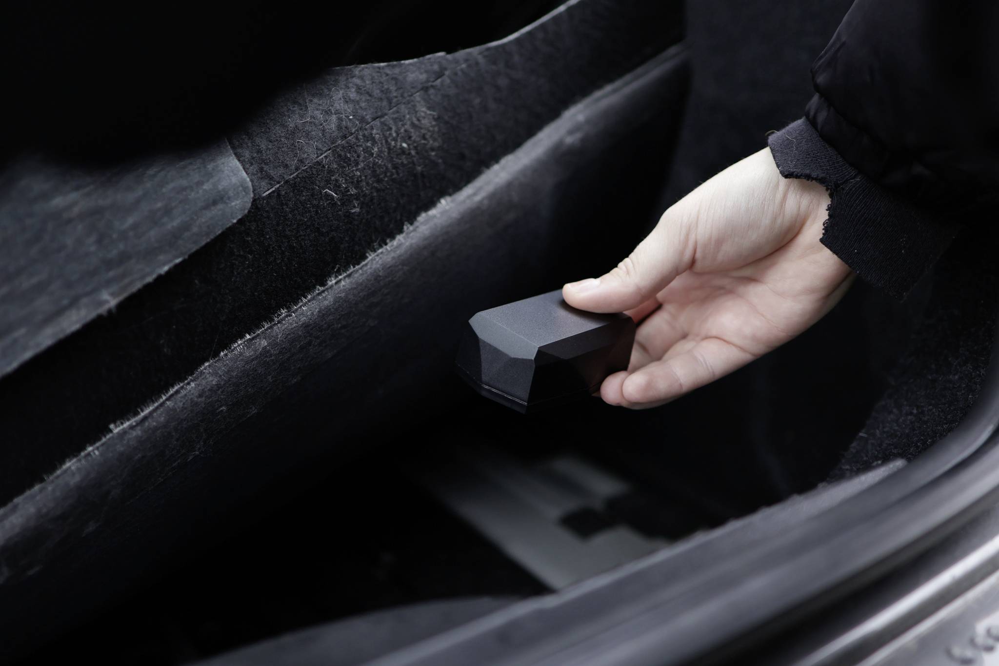 Een hand houdt een klein zwart voorwerp vast in de kofferbak van een auto. Het voorwerp lijkt een accessoire of gereedschap te zijn.