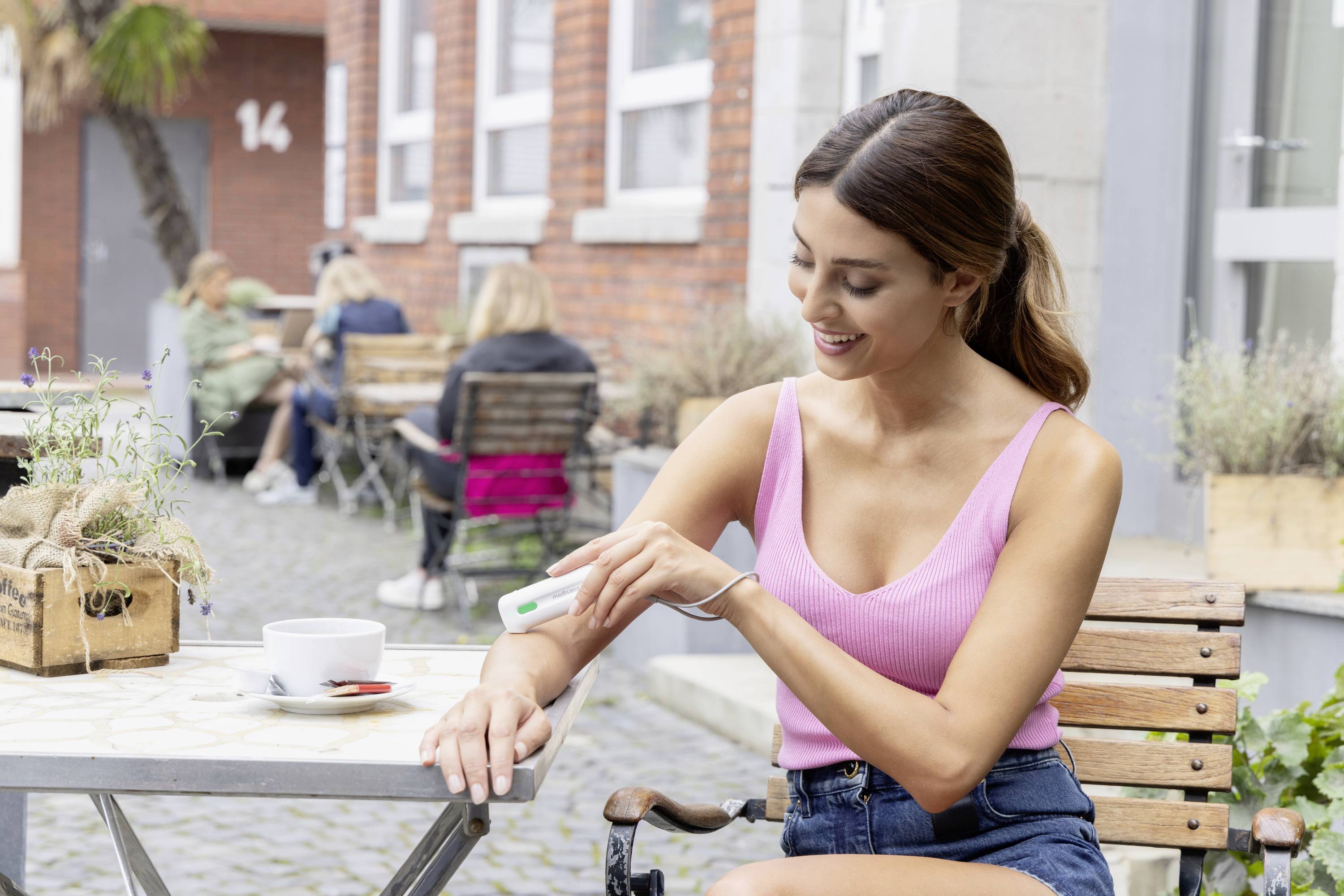 Een vrouw zit buiten aan een tafel en controleert haar arm met een apparaat. Op tafel staat een kopje koffie.