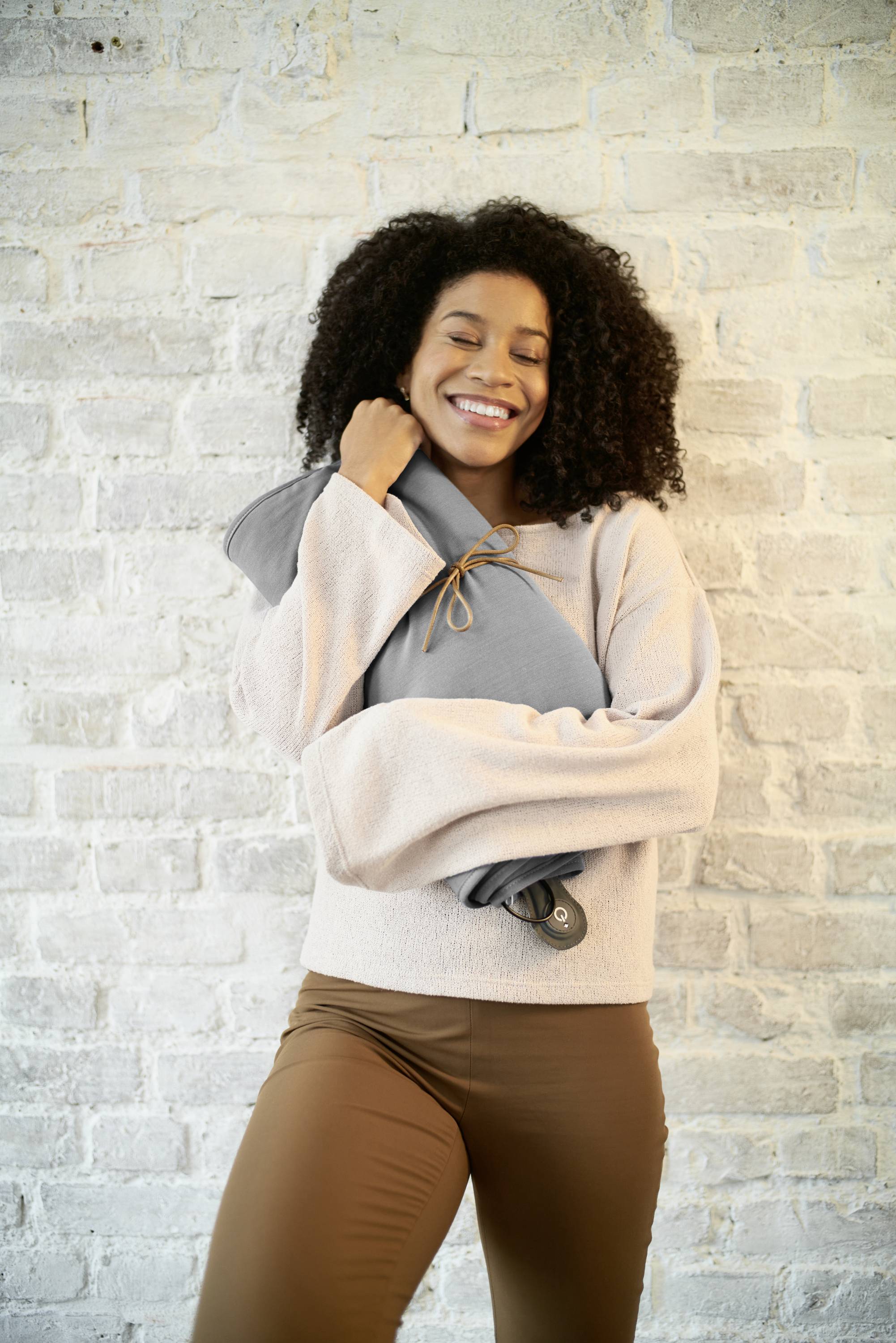 Vrouw in beige kleding glimlacht en houdt een warm, grijs kussen voor een witte bakstenen muur.