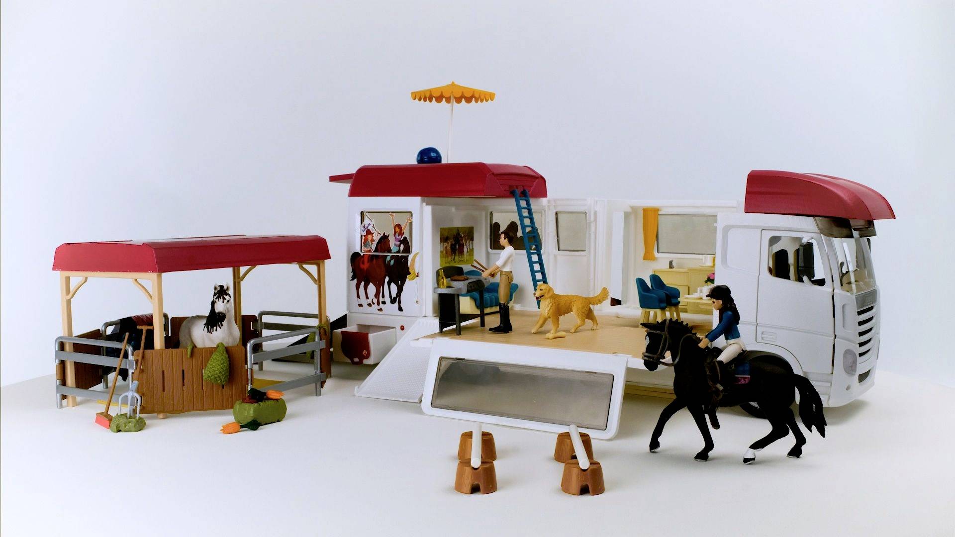 Een grote speelgoed-vrachtwagen met uitklapbare zijkanten toont een woonkamer en een terras. Figuren en een paard bevinden zich in de buurt.