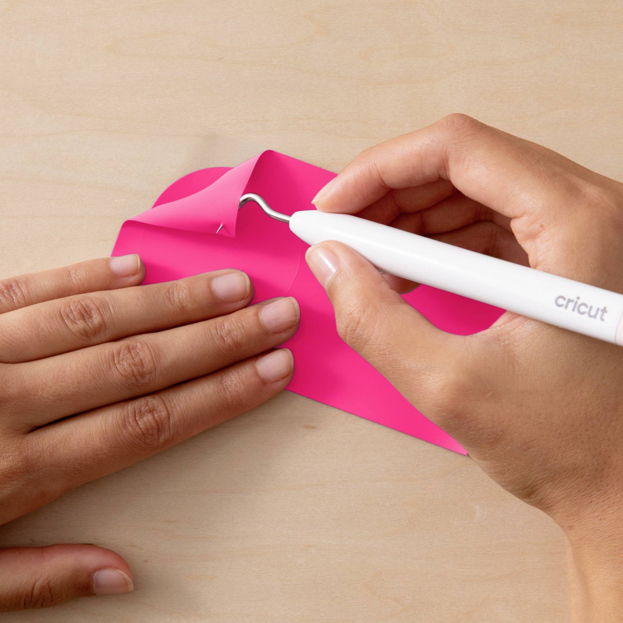 Twee handen verwijderen een roze vinylfolie van hout. Eén hand houdt een witte Cricut-ontgitterpen vast, terwijl de andere hand de folie vasthoudt.