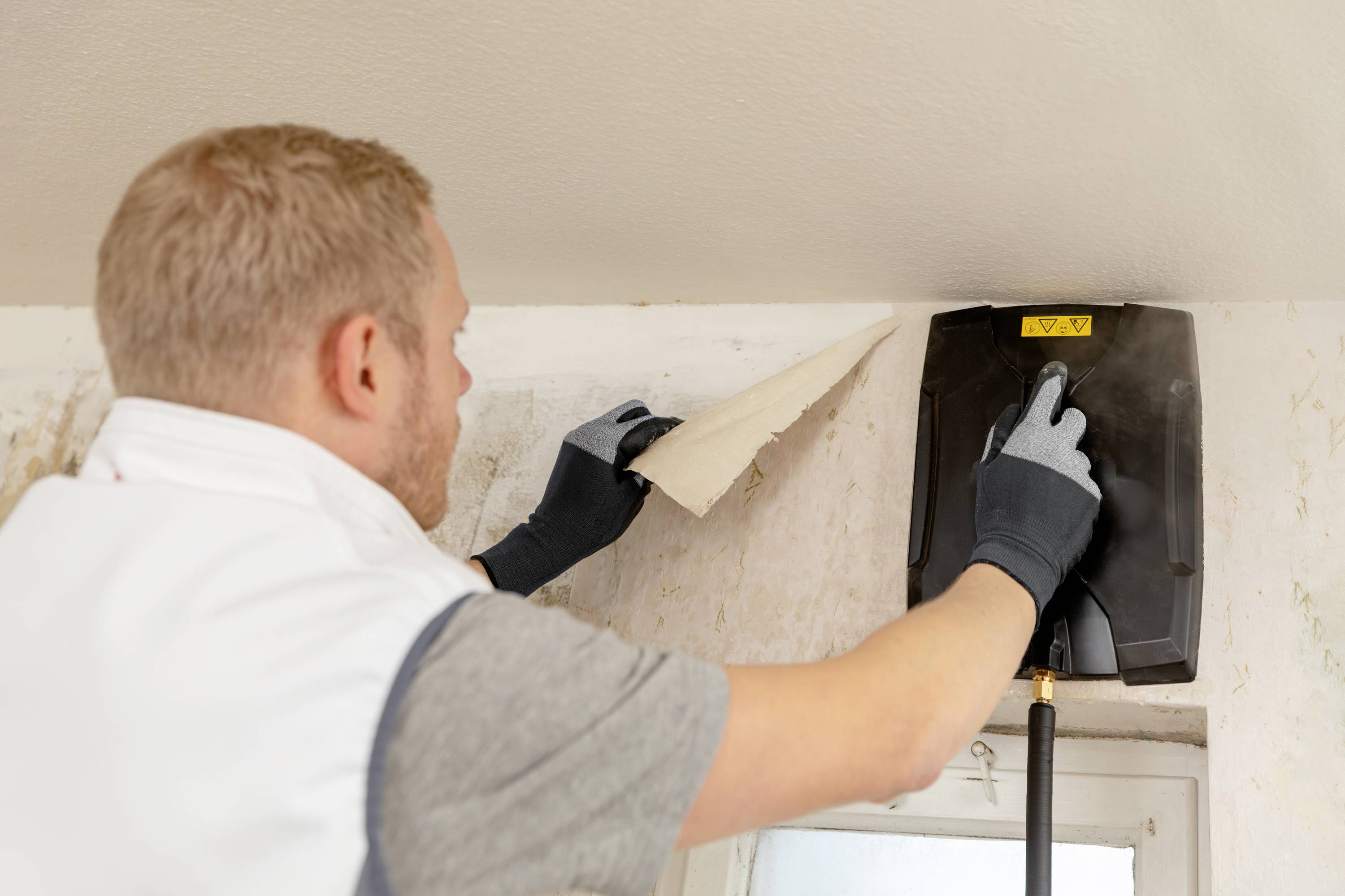 Een man verwijdert behang van een plafond met behulp van een stoomreiniger. Hij draagt handschoenen en is geconcentreerd aan het werk.