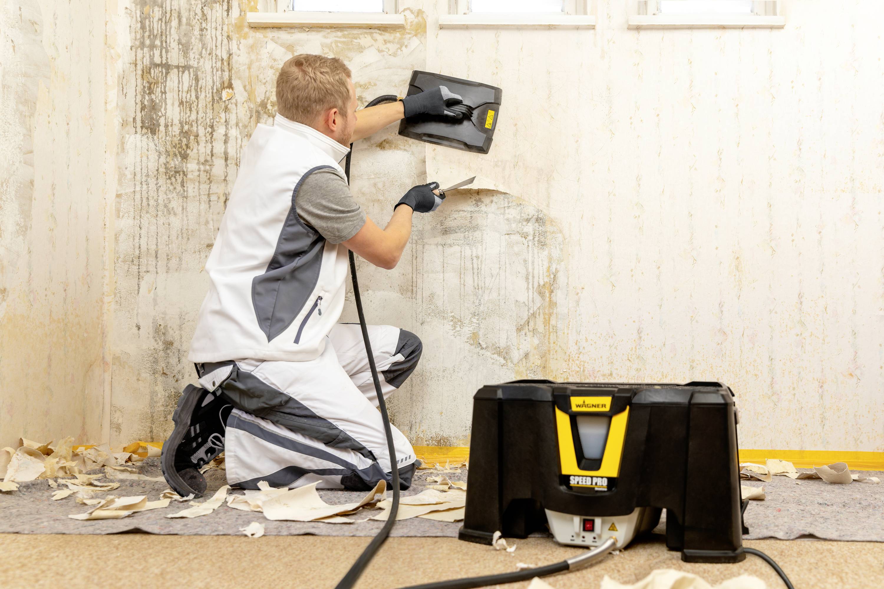 Een man in werkkleding verwijdert behang van een muur met een behangstomer. Stukjes behang liggen op de vloer.