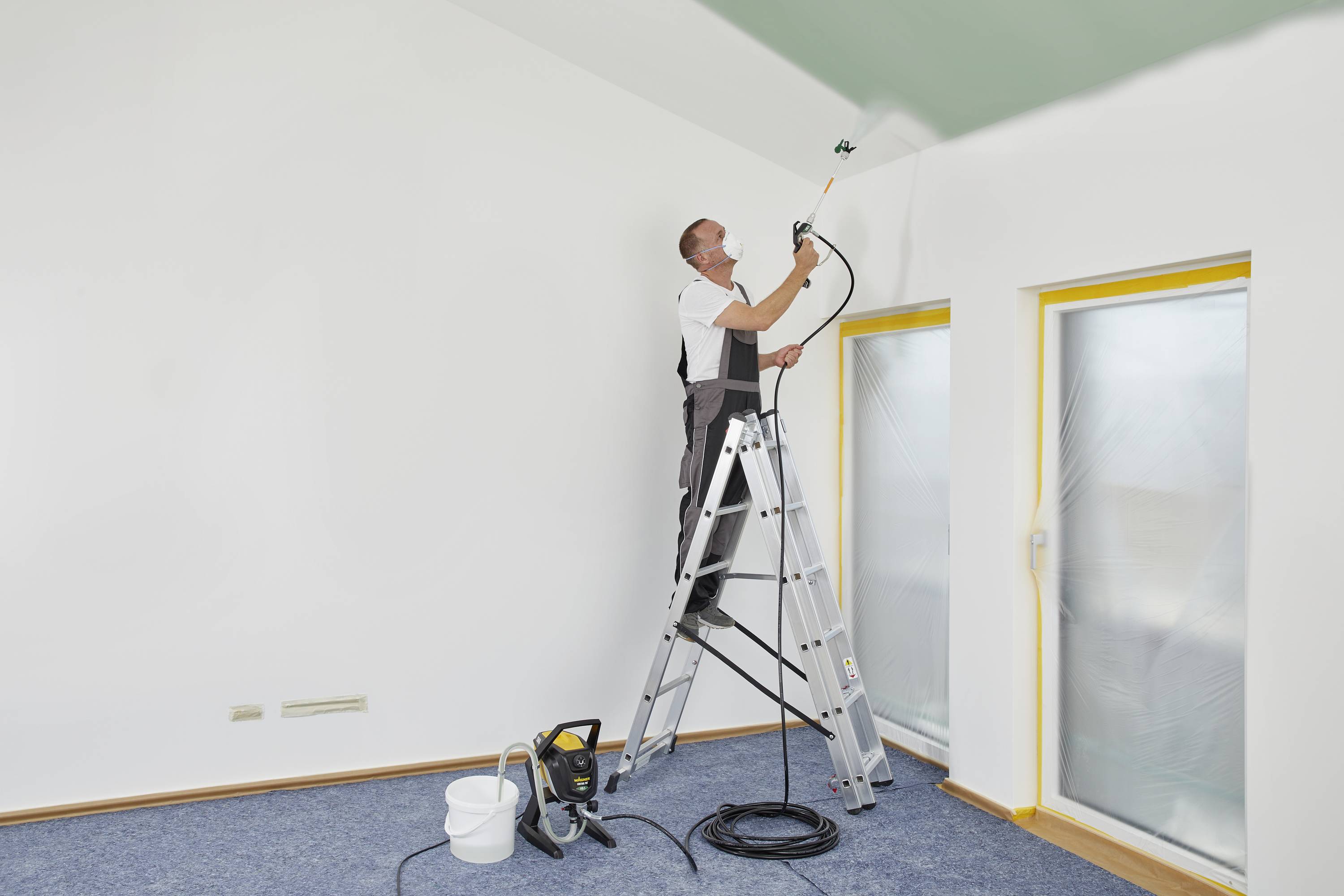 Een man staat op een ladder en schildert het plafond van een kamer met groene verf. De raamruiten zijn afgedekt met folie.