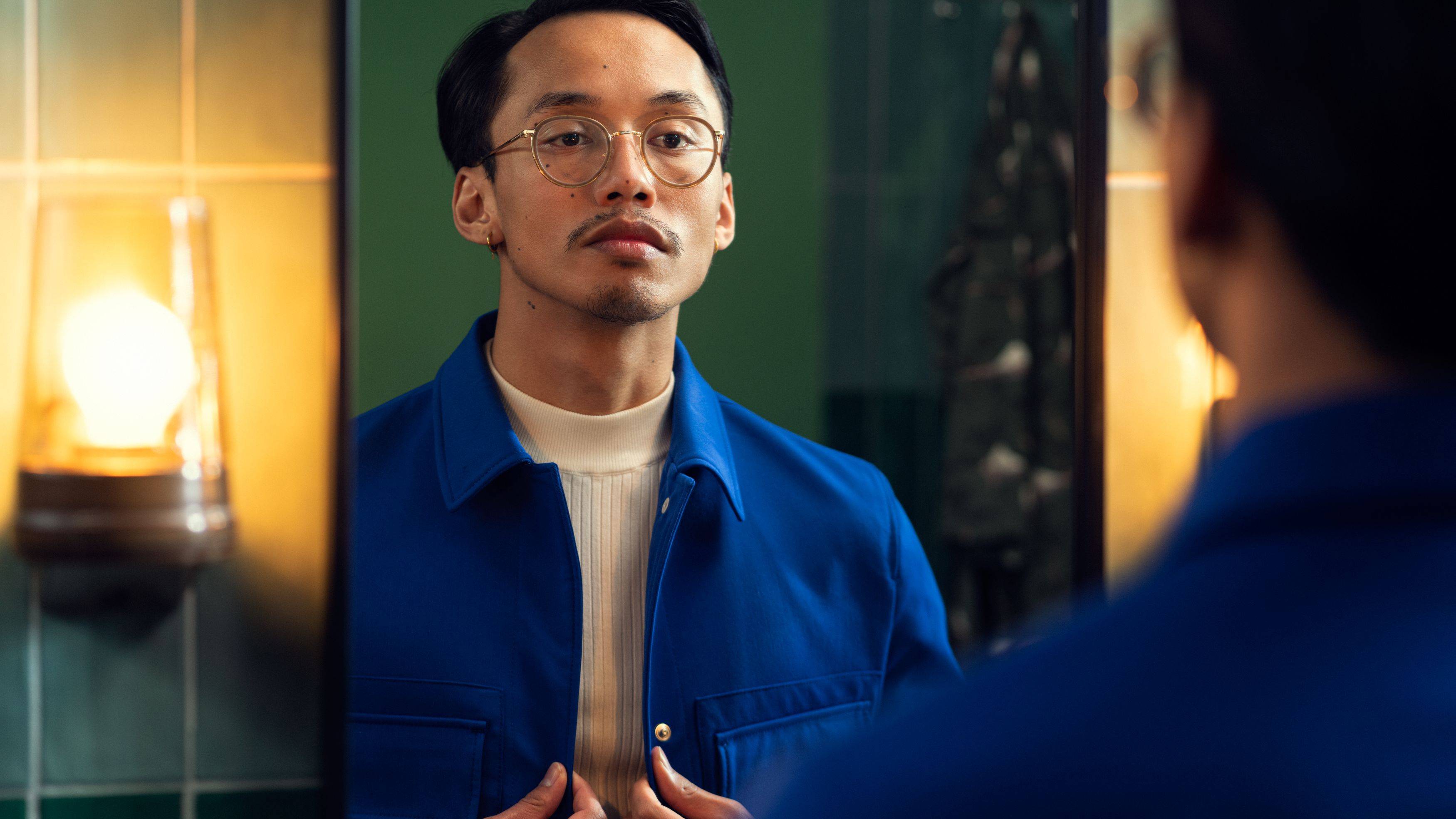 Een man in een blauw shirt kijkt naar zichzelf in een spiegel. Rechts in het beeld schijnt een lamp.