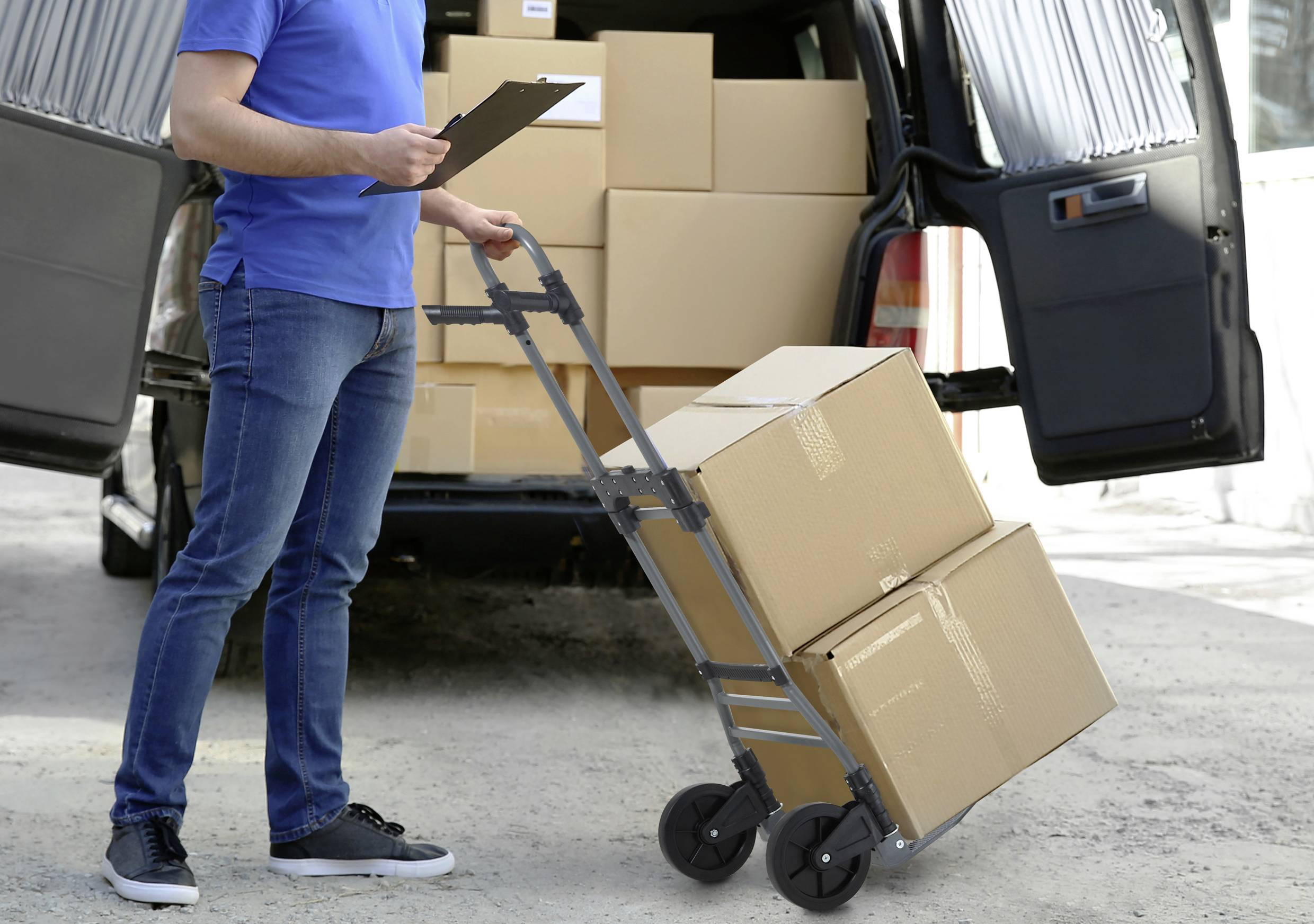 Een persoon in blauwe kleding gebruikt een steekwagen om dozen uit een open bestelwagen te lossen en maakt aantekeningen op een klembord.