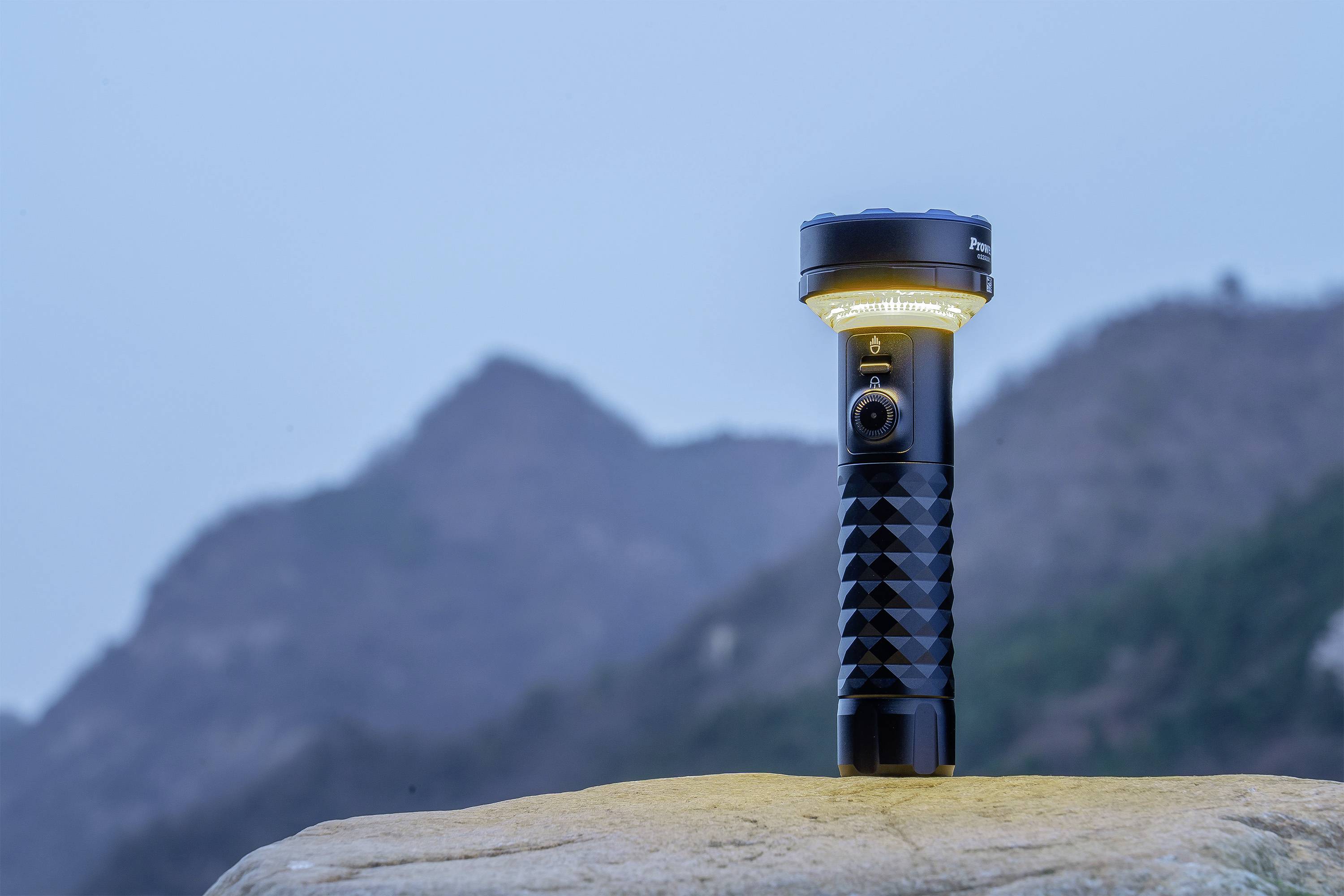 'Zaklamp op een rots voor een bergachtergrond bij schemering. De lamp schijnt en steekt af tegen de blauwe lucht.'