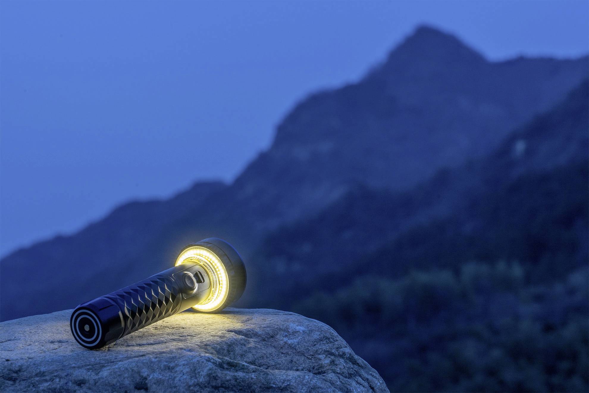 Een oplichtende zaklamp ligt op een rots in een bergachtig landschap bij schemering. De omgeving is donkerblauw.