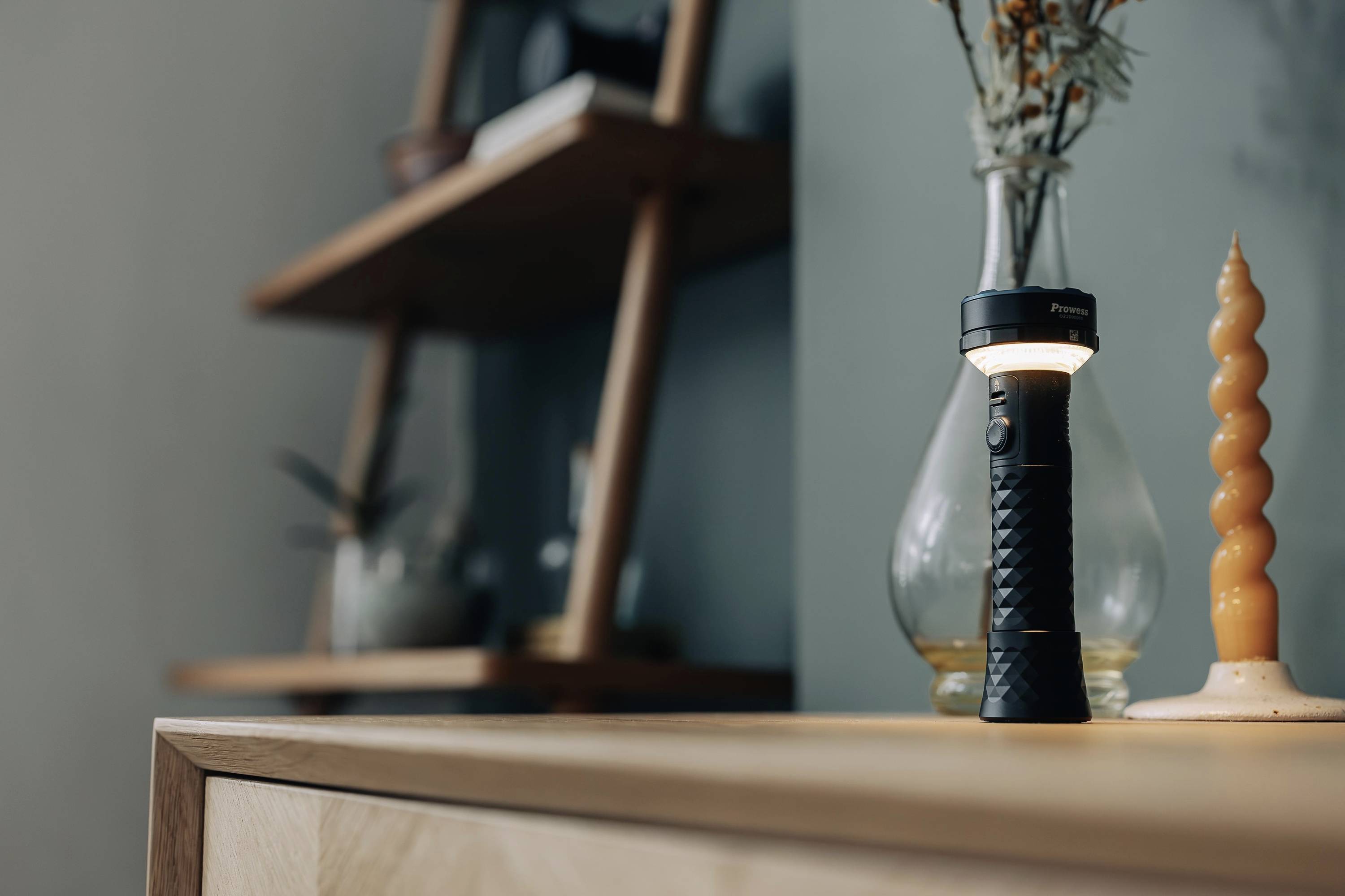 Een zaklamp staat ingeschakeld op een tafel naast een gedraaide kaars en een glazen voorwerp, op de achtergrond een rek.