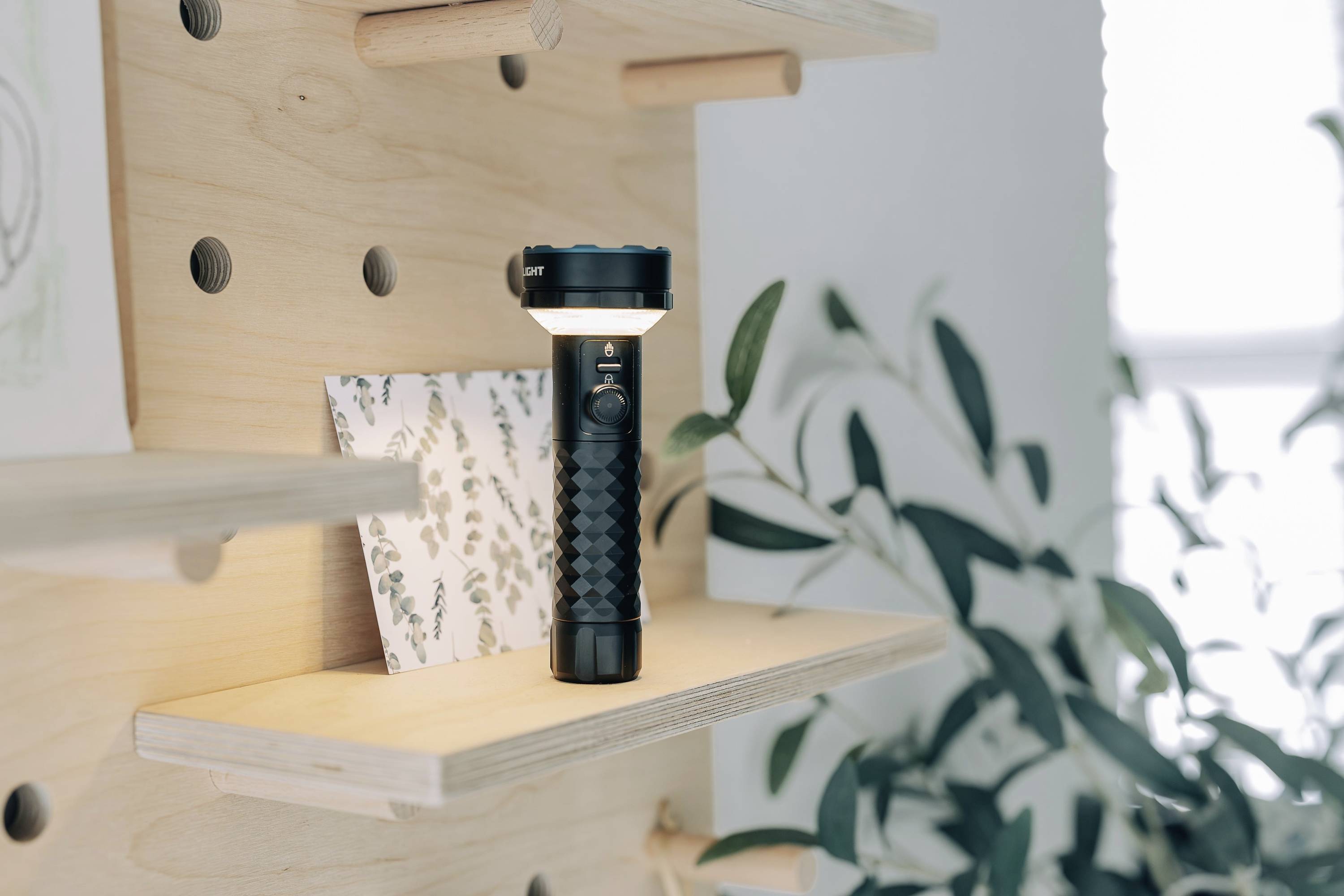 Een zwarte zaklamp staat op een houten rek, omgeven door planten en een blad met plantaardige patronen.