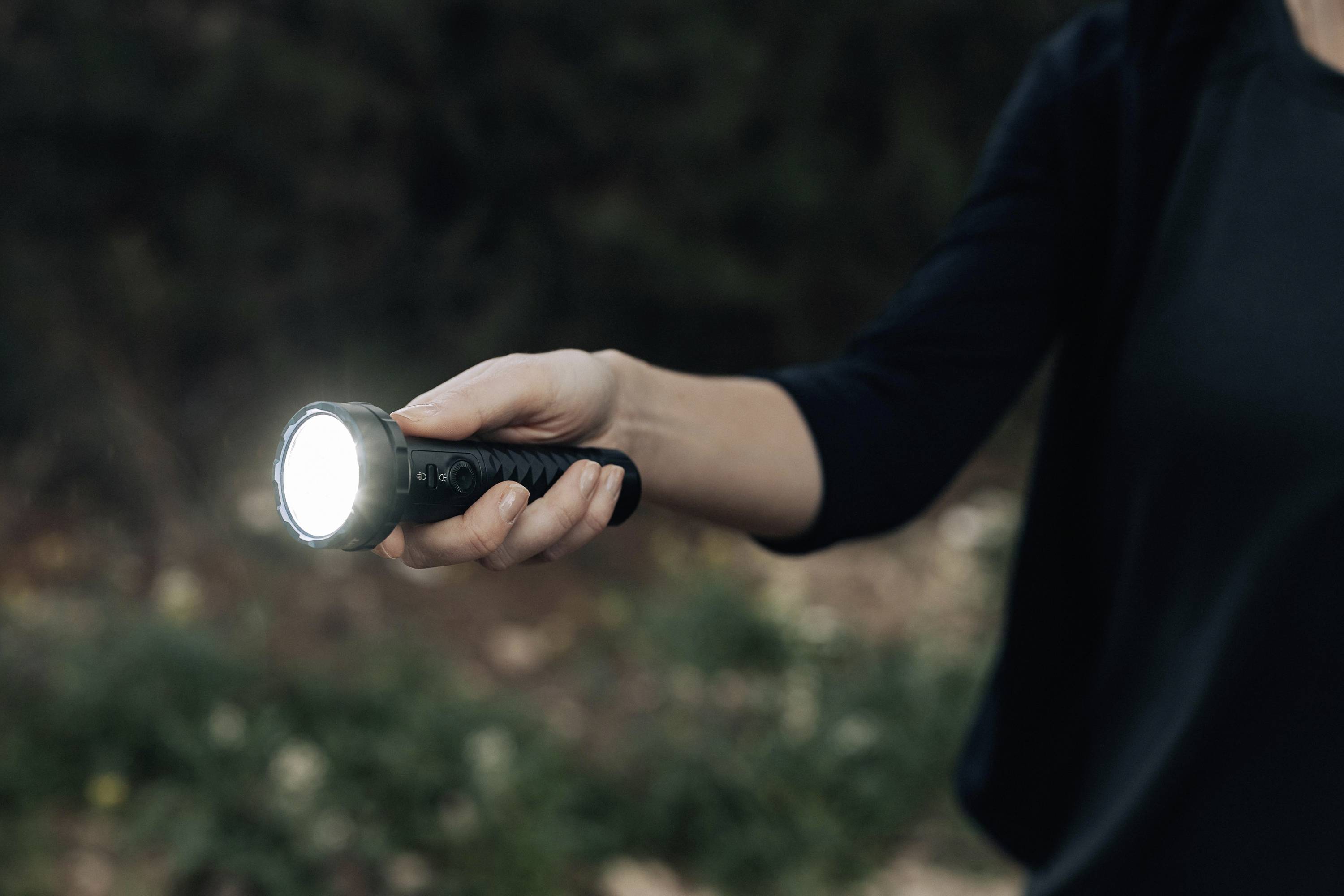 Een persoon houdt een brandende zaklamp vast. De achtergrond is wazig en lijkt op een bos of tuin.