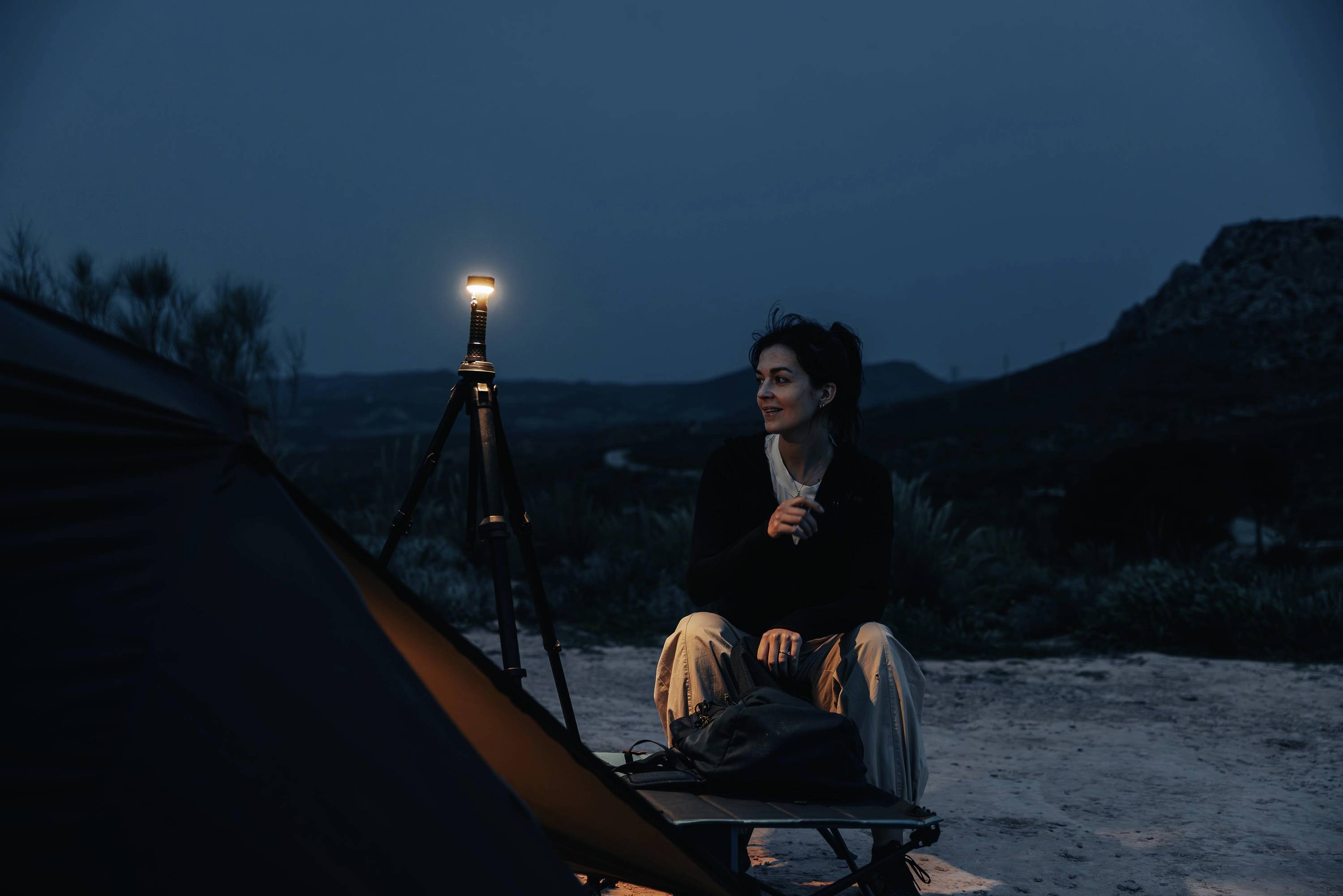 Een vrouw zit in het donker bij een lamp naast een tent in een landschap met heuvels, ogenschijnlijk verzonken in gedachten.