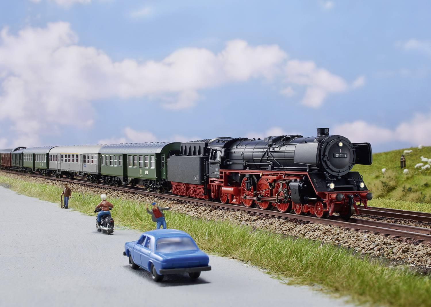 Een zwarte stoomtrein rijdt over de rails door een groene landschap onder een blauwe lucht. Vooraan in beeld zijn een auto en mensen te zien.