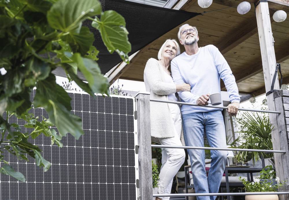Een ouder echtpaar staat op een terras naast zonnepanelen. Ze kijken samen in de verte en zien er tevreden en ontspannen uit.