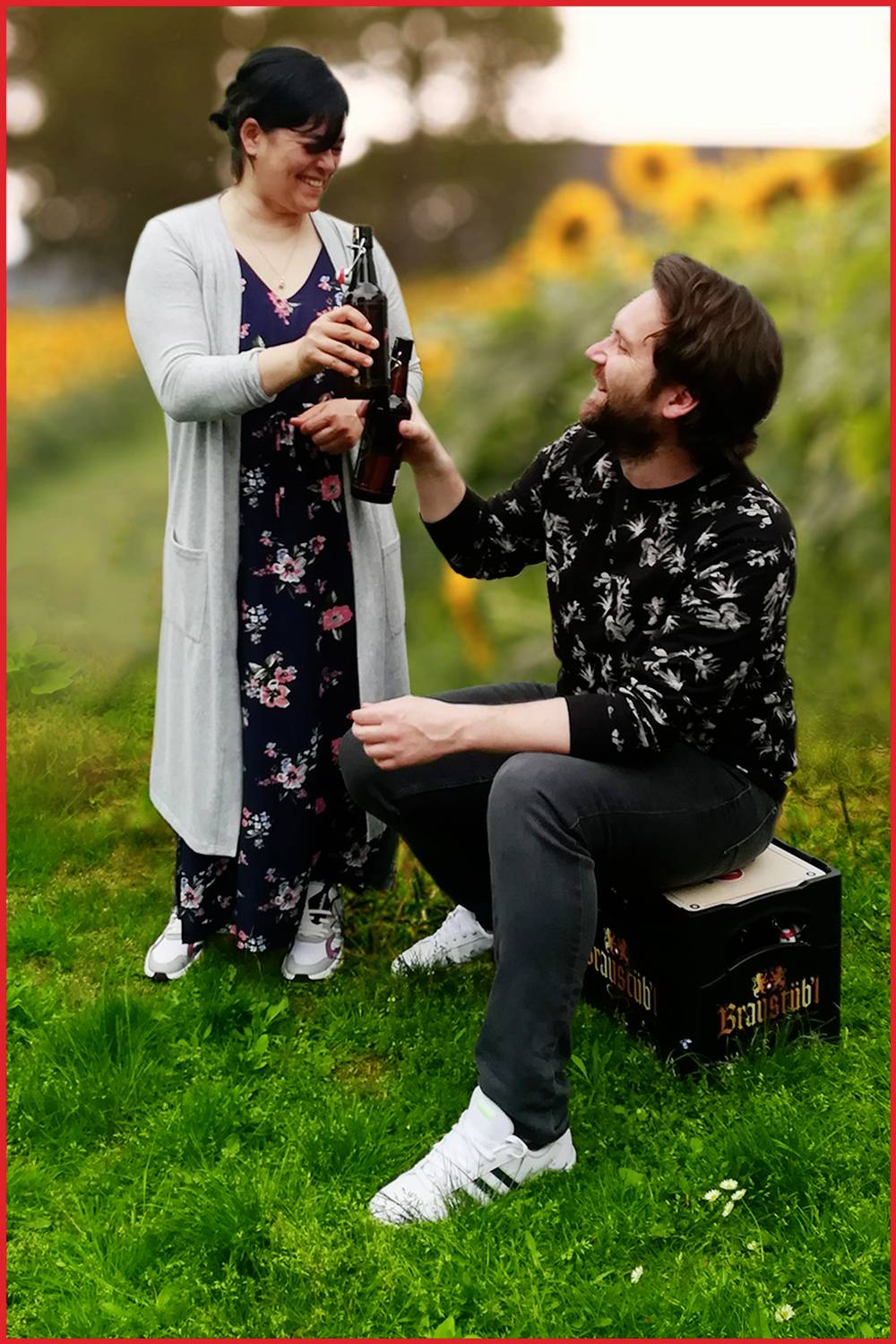 Een man zit op een bierkrat terwijl een vrouw staat. Beiden hebben een fles in de hand en heffen het glas op elkaar, omringd door zonnebloemen.