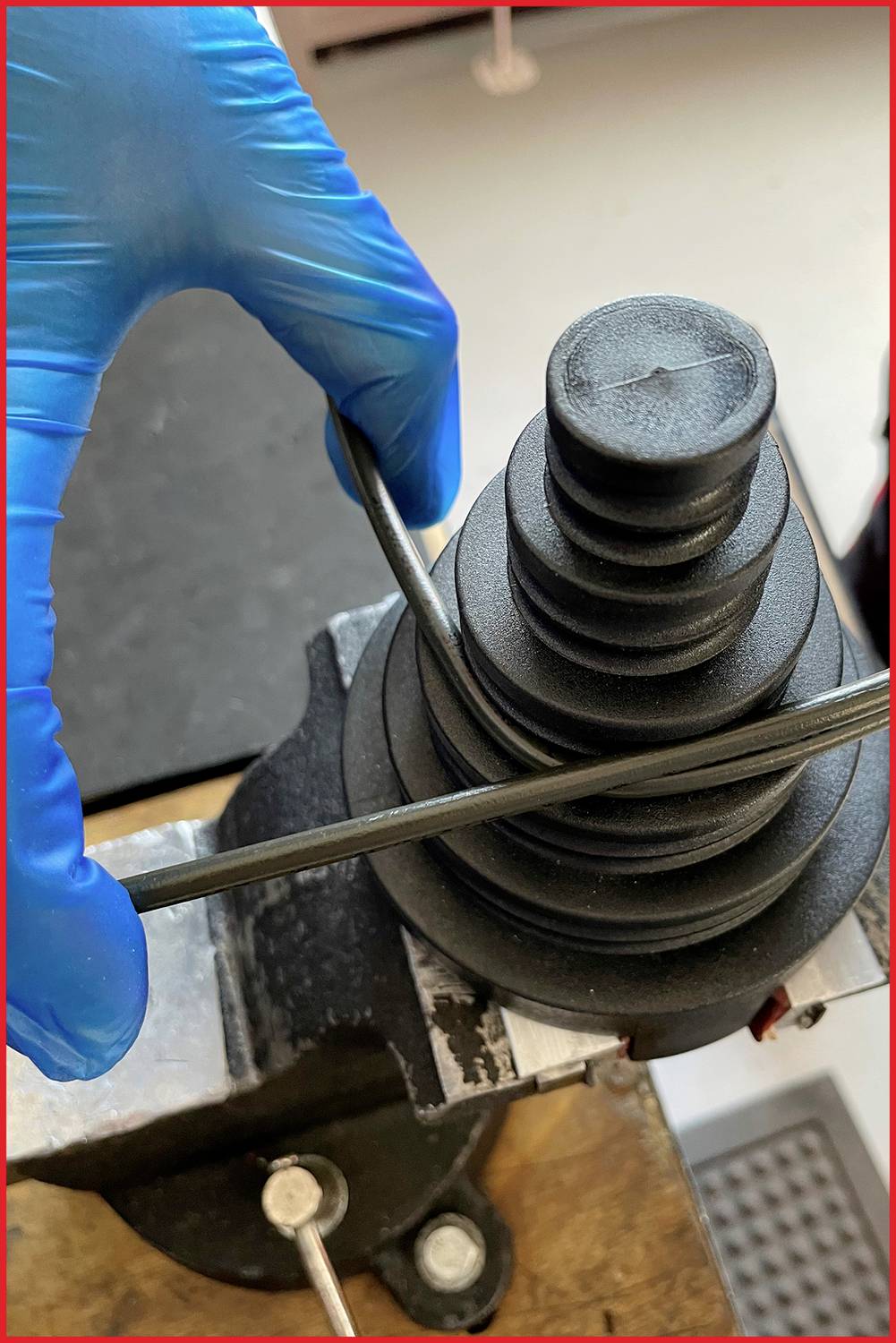 Een persoon draagt blauwe beschermende handschoenen en buigt een metalen onderdeel met een bankschroef en een cilindrisch gereedschap.