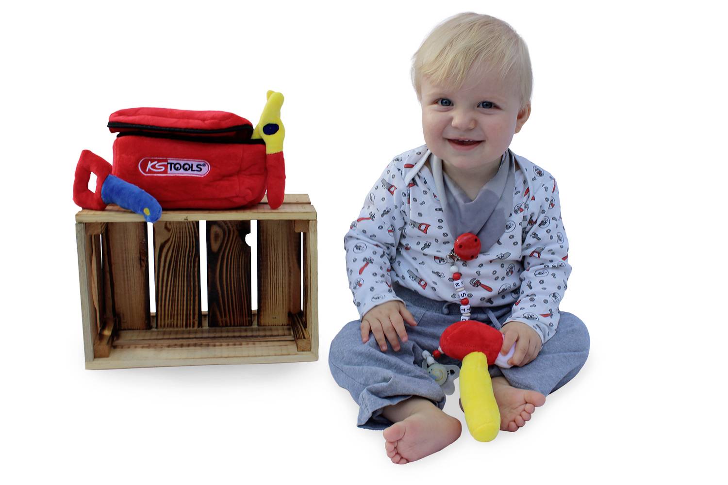 Een peuter zit lachend naast een stoffen gereedschapskist op een houten kist met een speelgoedmoker.