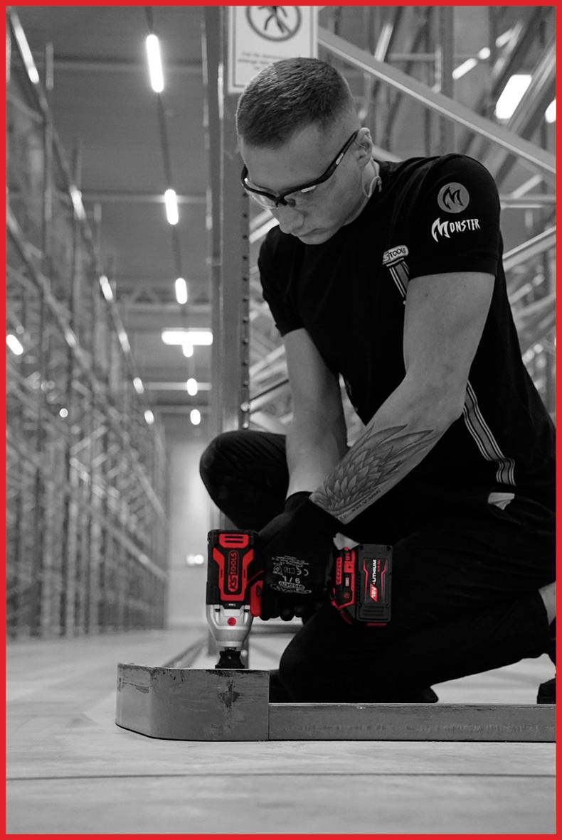 Man in werkkleding knielt op de magazijnvloer en bevestigt een metalen onderdeel met een accuschroefmachine. Op de achtergrond zijn stellingen zichtbaar.