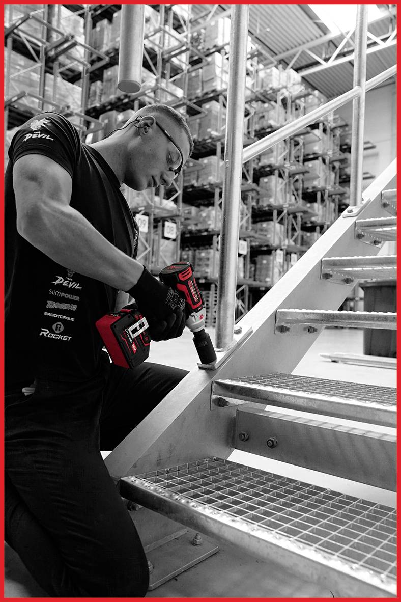 Een man monteert met een accuschroefmachine op een metalen trap in een magazijn. Op de achtergrond zijn rekken met dozen zichtbaar.