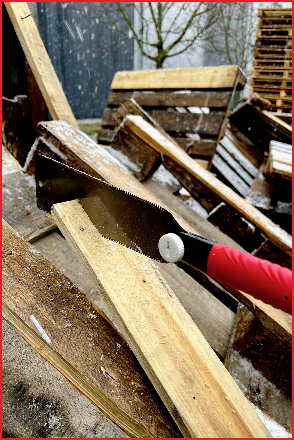 Een handzaag met een rode handgreep zaagt een houten plank. Op de achtergrond zijn houten planken gestapeld. De omgeving heeft een winterlijke sfeer.