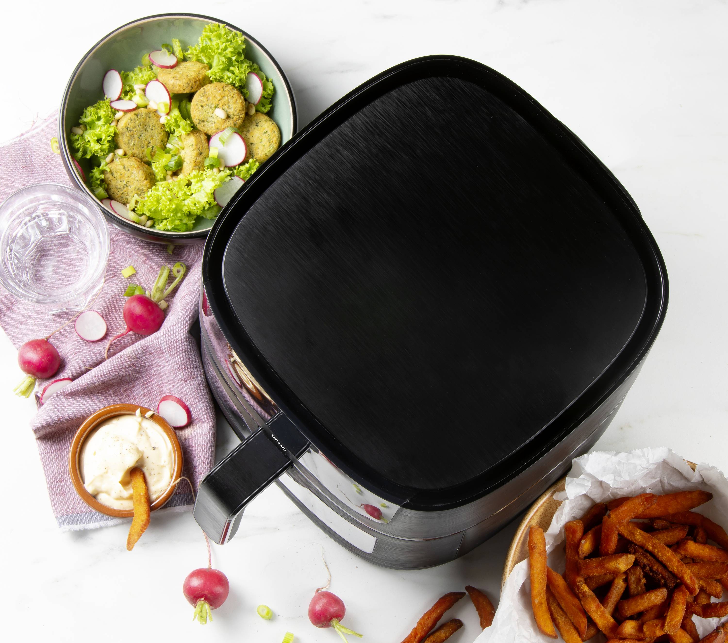 Een airfryer staat op een tafel. Ernaast staat een bord met falafel en groenten, een kom zoete aardappelfrieten en een dip.