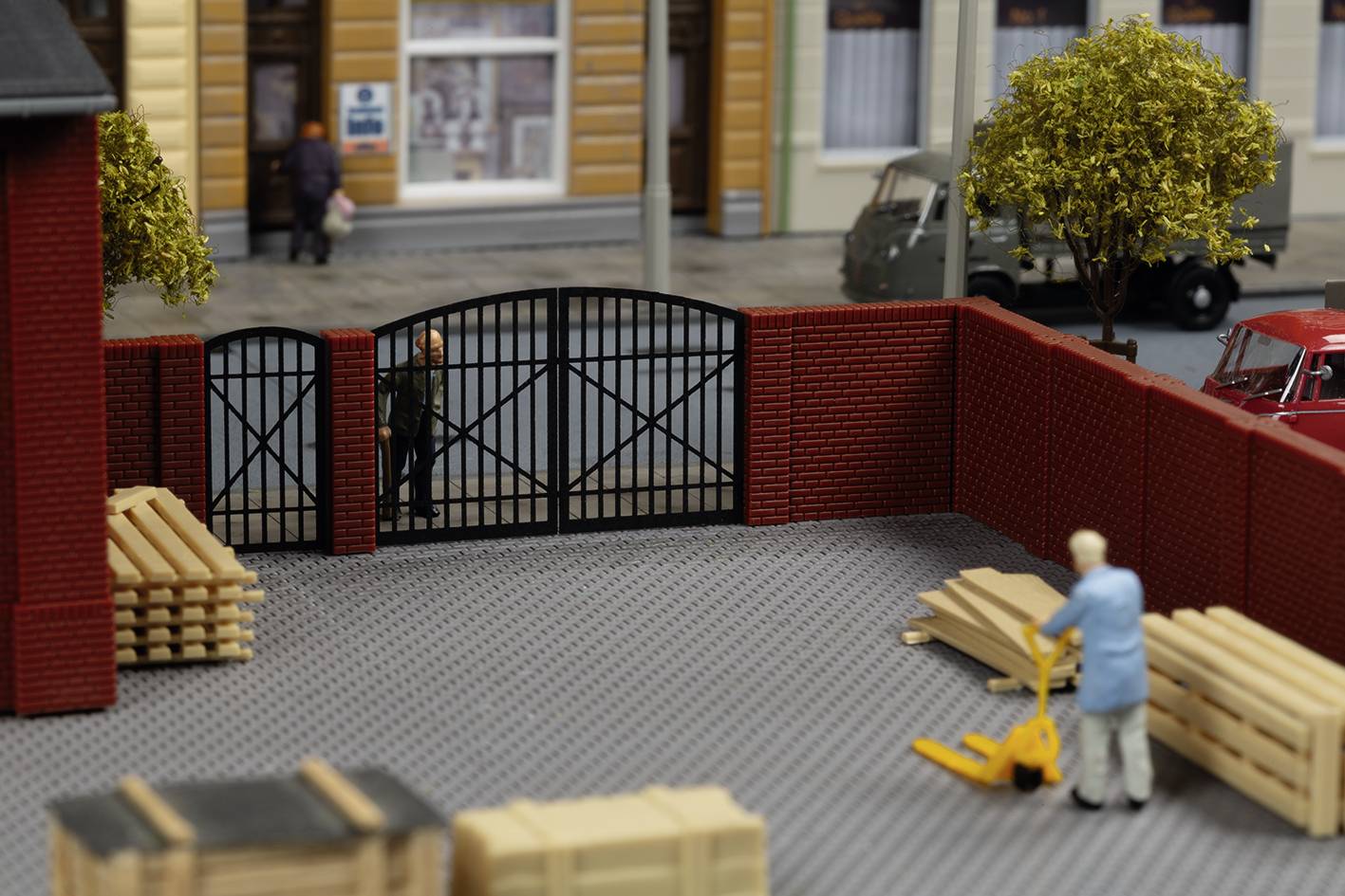 Een model van een stedelijke scene: Een man met een pompwagen verplaatst pallets in een omheinde binnenplaats; op de achtergrond lopen voorbijgangers.