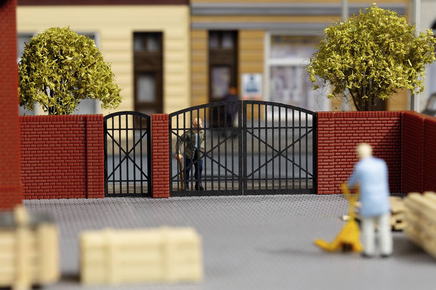 Een miniatuurscène toont een poort voor een gebouw met twee bomen, een man op de achtergrond en een persoon op de voorgrond die een pallet verplaatst.