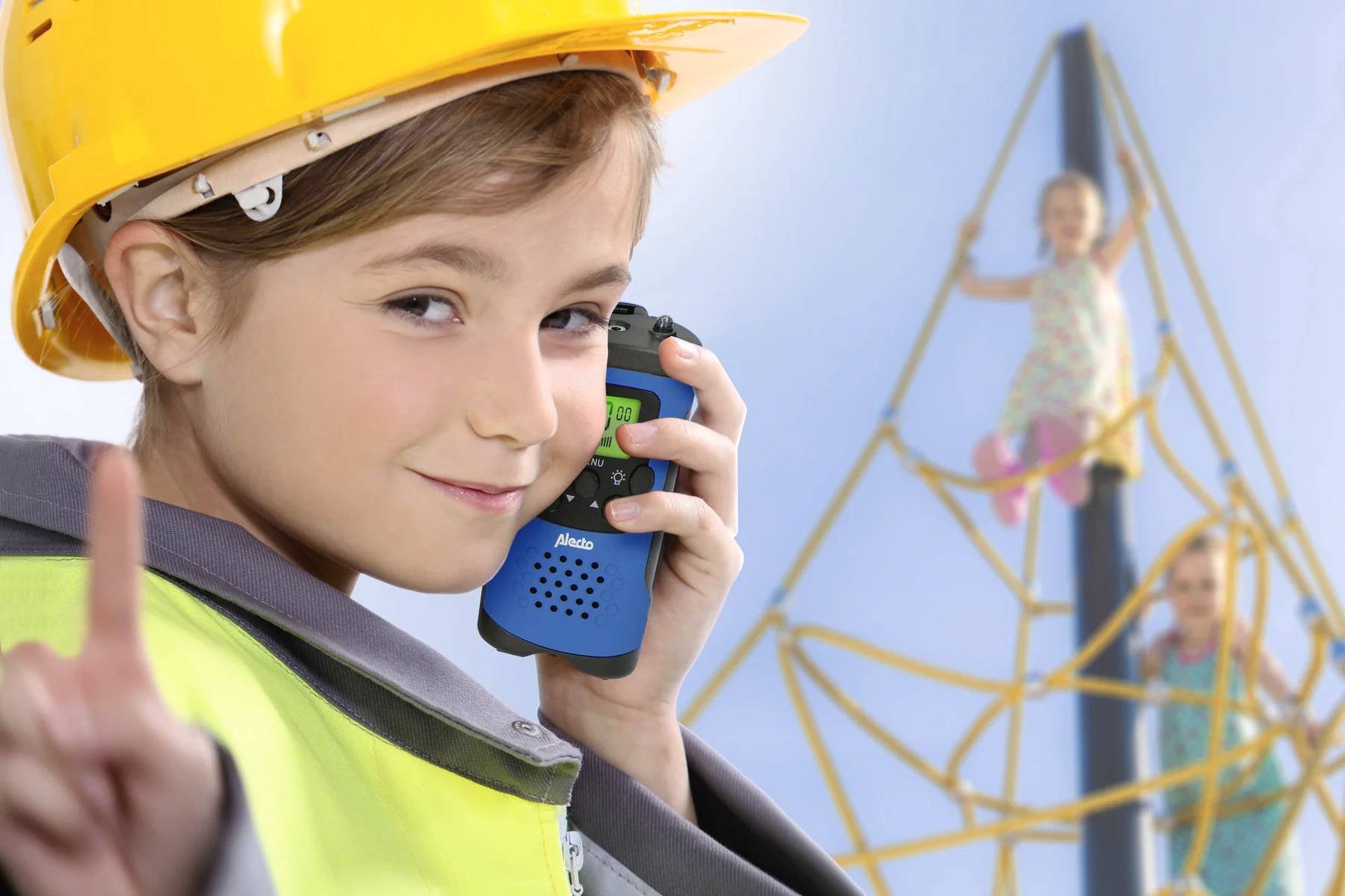 Een kind draagt een bouwveiligheidshelm en houdt een walkietalkie vast, terwijl twee kinderen op de achtergrond op een klimrek spelen.